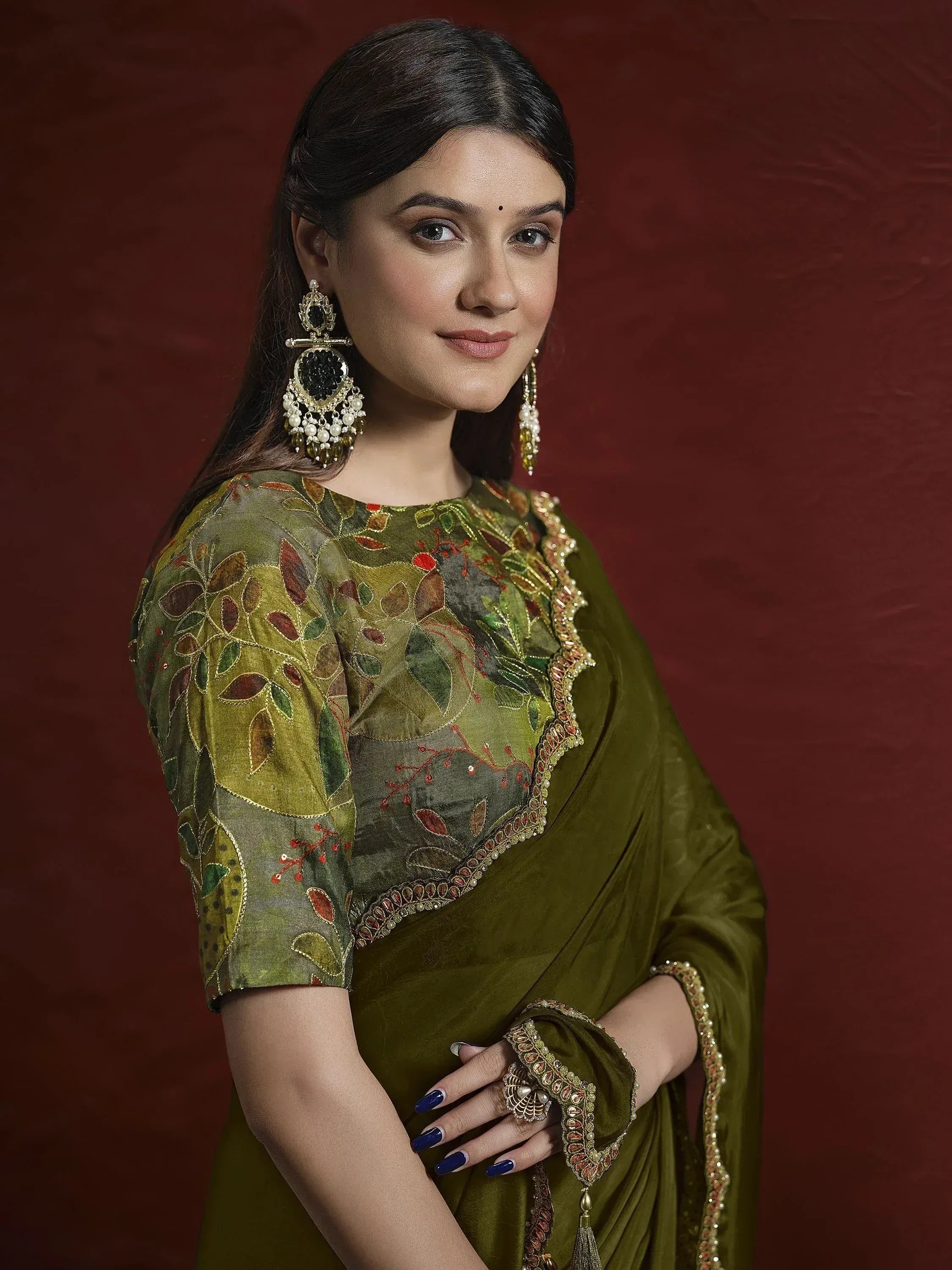 Woman in olive green embroidered saree with floral blouse, gold jewelry, and blue nails against maroon backdrop