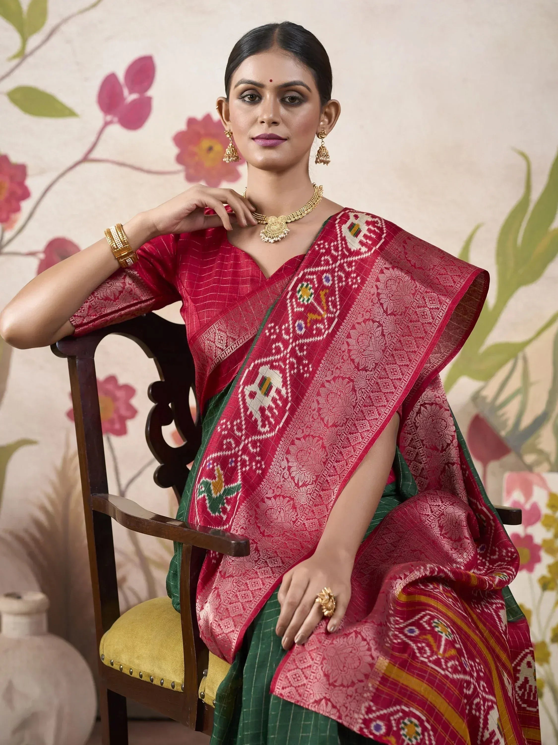 Woman in red and green ethnic saree with elephant motifs, gold jewelry, seated on wooden chair