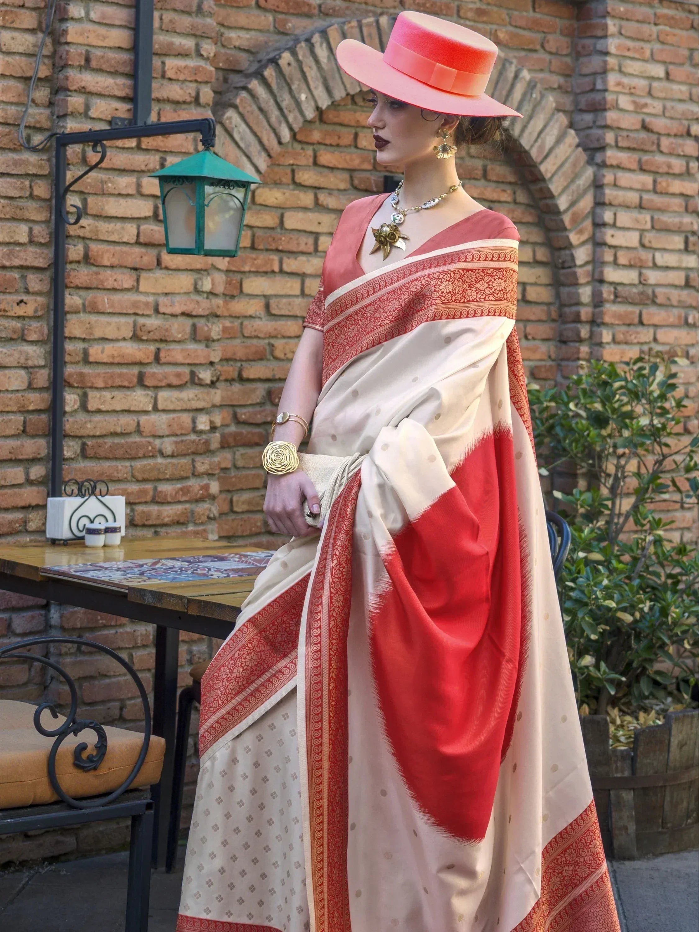 Woman in elegant cream and red saree with intricate border, styled with pink hat and gold jewelry, perfect for ethnic occasions.