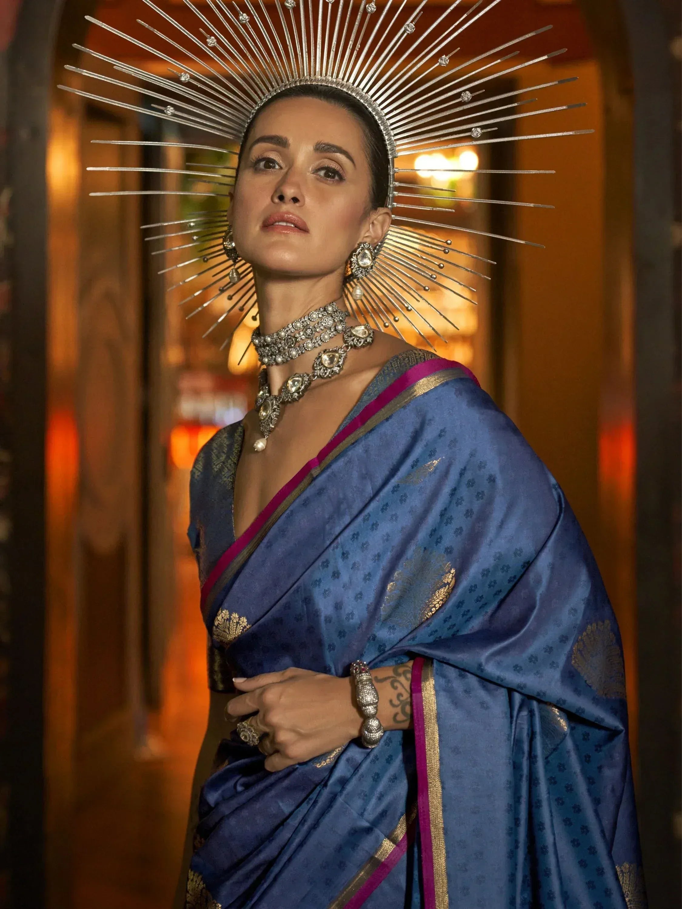 Woman in blue designer saree with gold motifs, statement sunburst headpiece, and ethnic jewelry