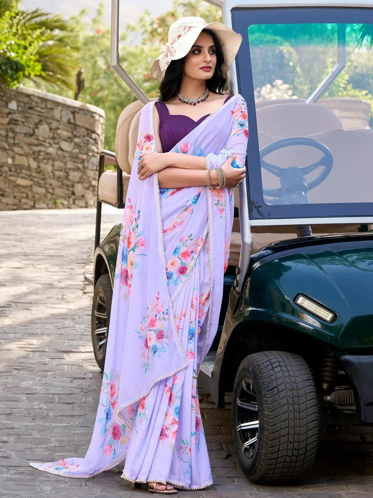 Woman in purple floral saree with matching blouse, standing beside golf cart, wearing floral hat and jewelry