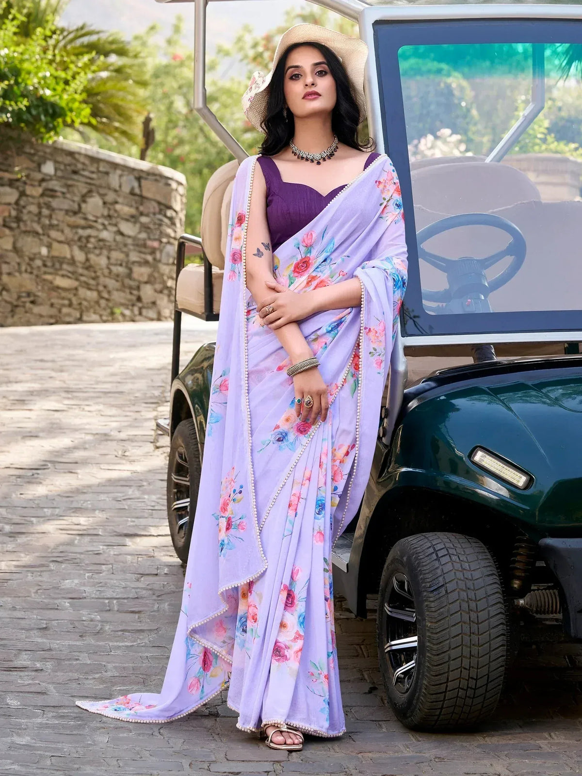 Woman in lavender floral saree with purple blouse, standing beside golf cart, stylish ethnic wear
