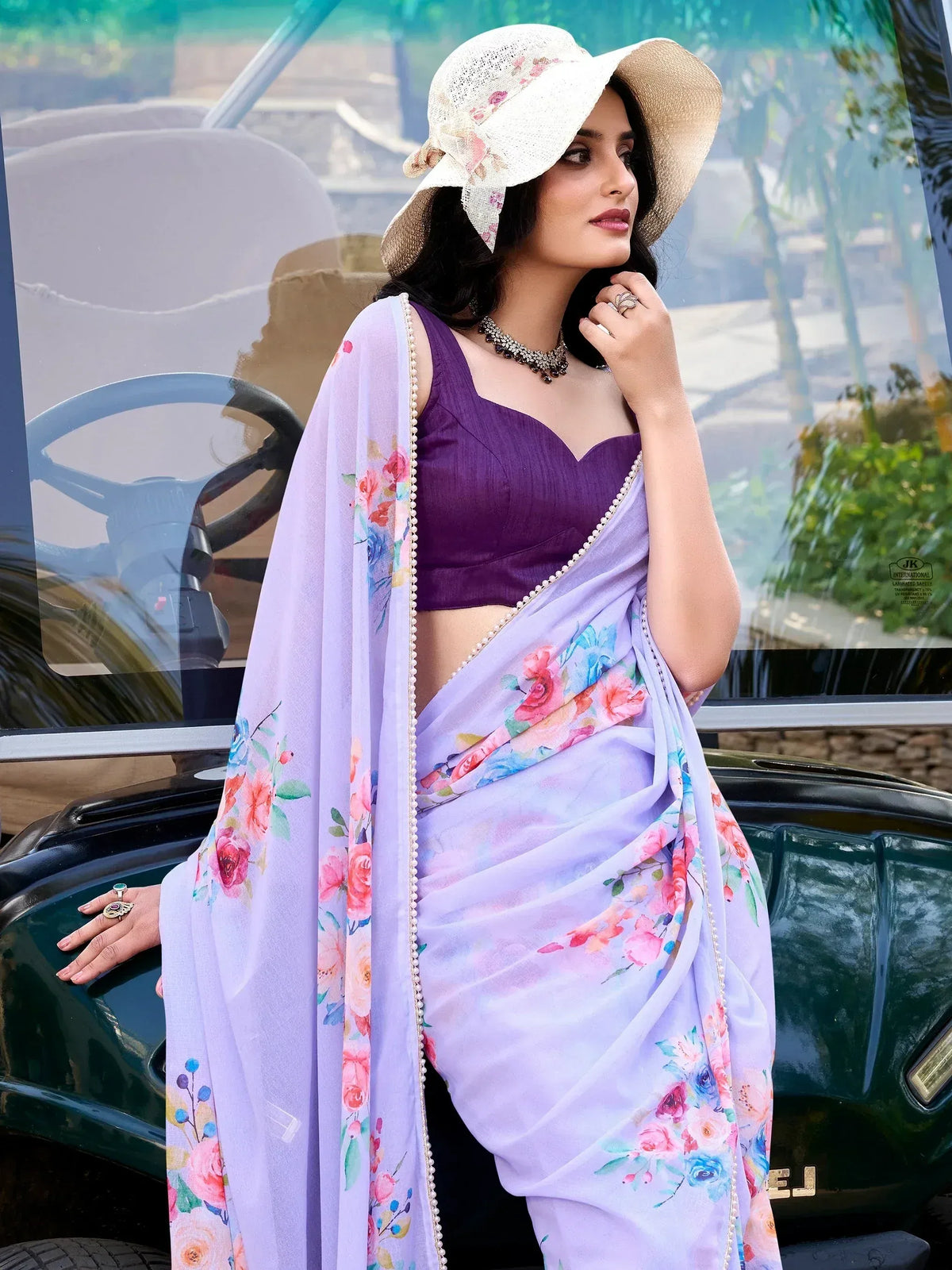 Woman in purple floral saree with pearl border, styled with wide-brim hat, standing beside vintage car