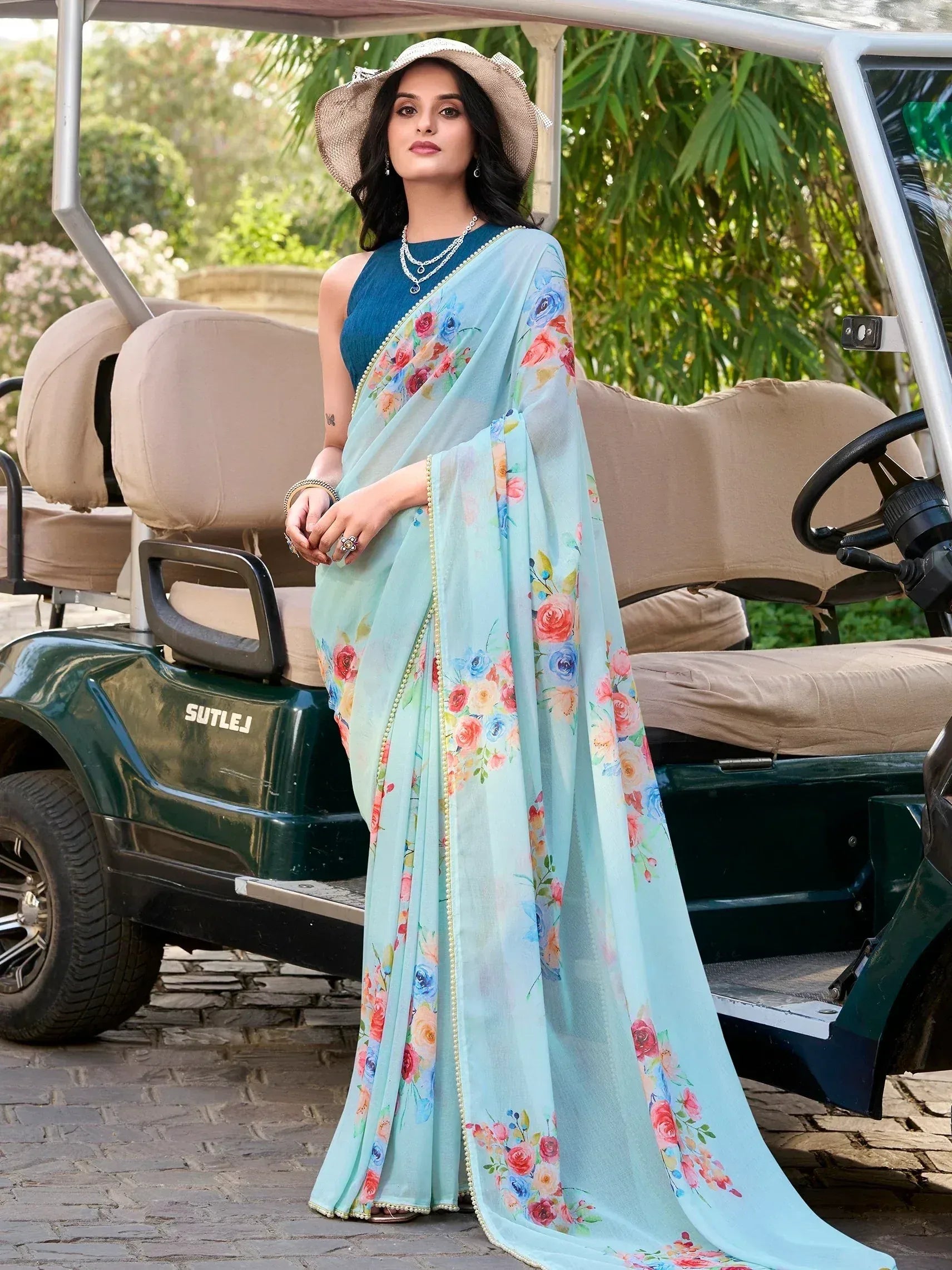 Woman in floral print light blue saree with blue blouse, standing beside golf cart, wearing straw hat