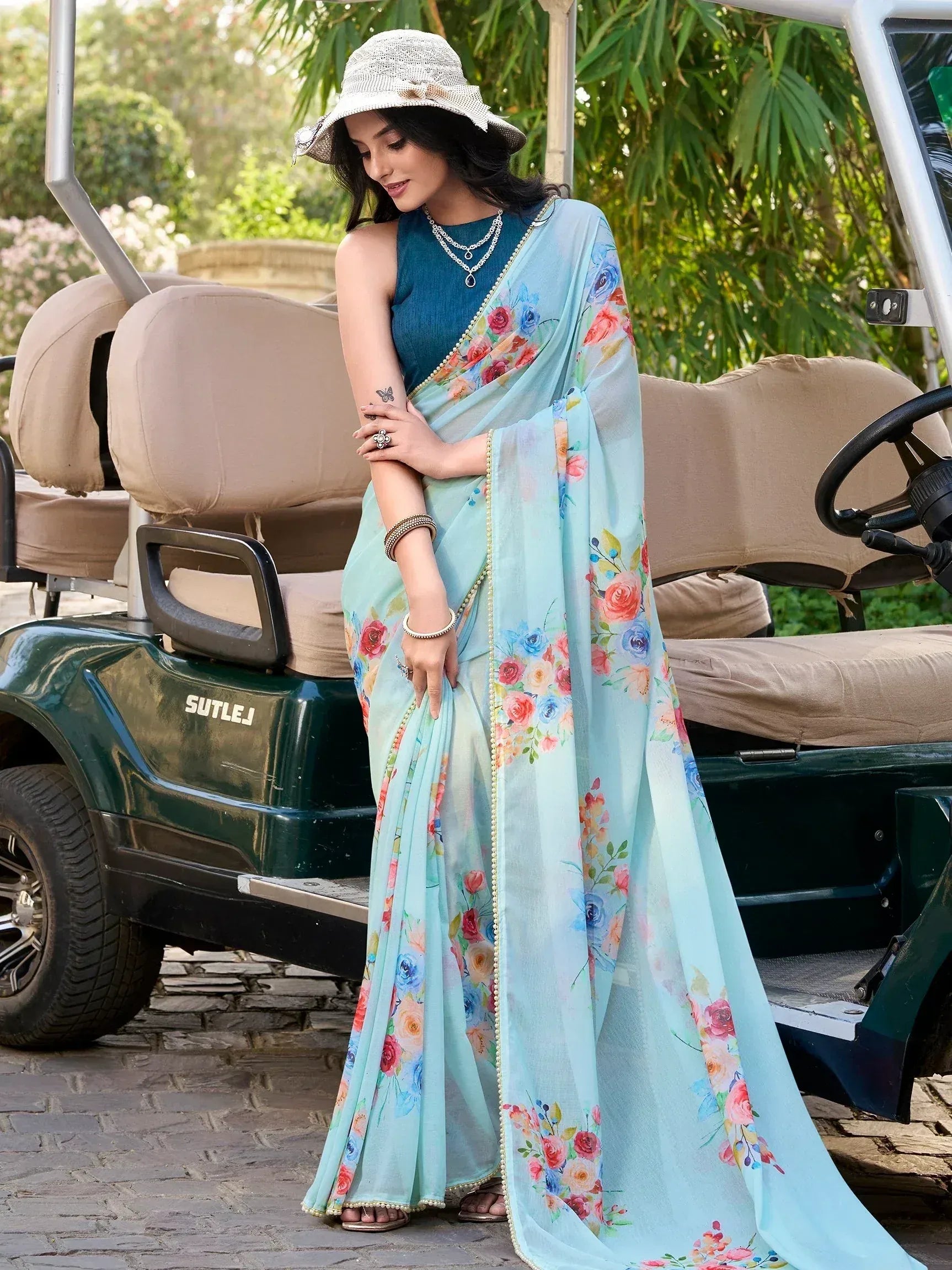 Woman in floral print light blue saree with gold trim, standing beside golf cart, wearing hat and jewelry