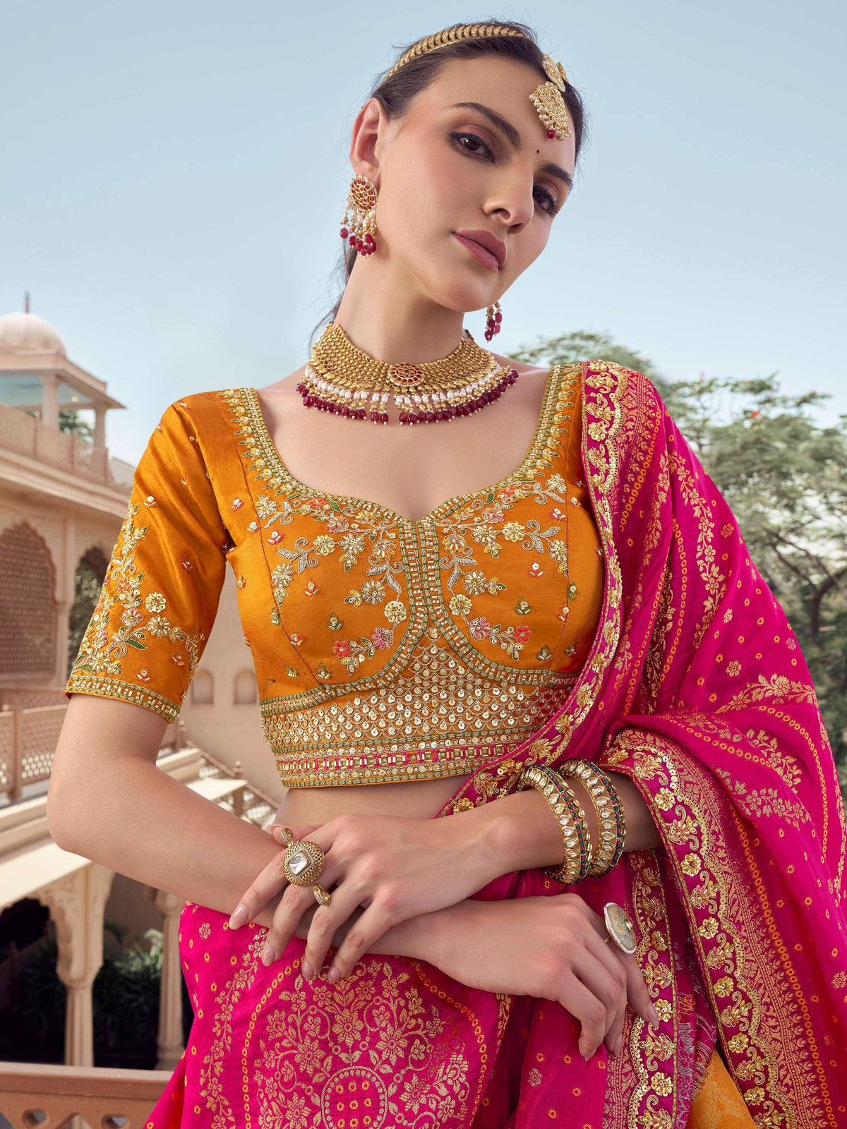 Woman in vibrant pink and gold embroidered lehenga choli with traditional Indian jewelry, perfect for festive occasions.