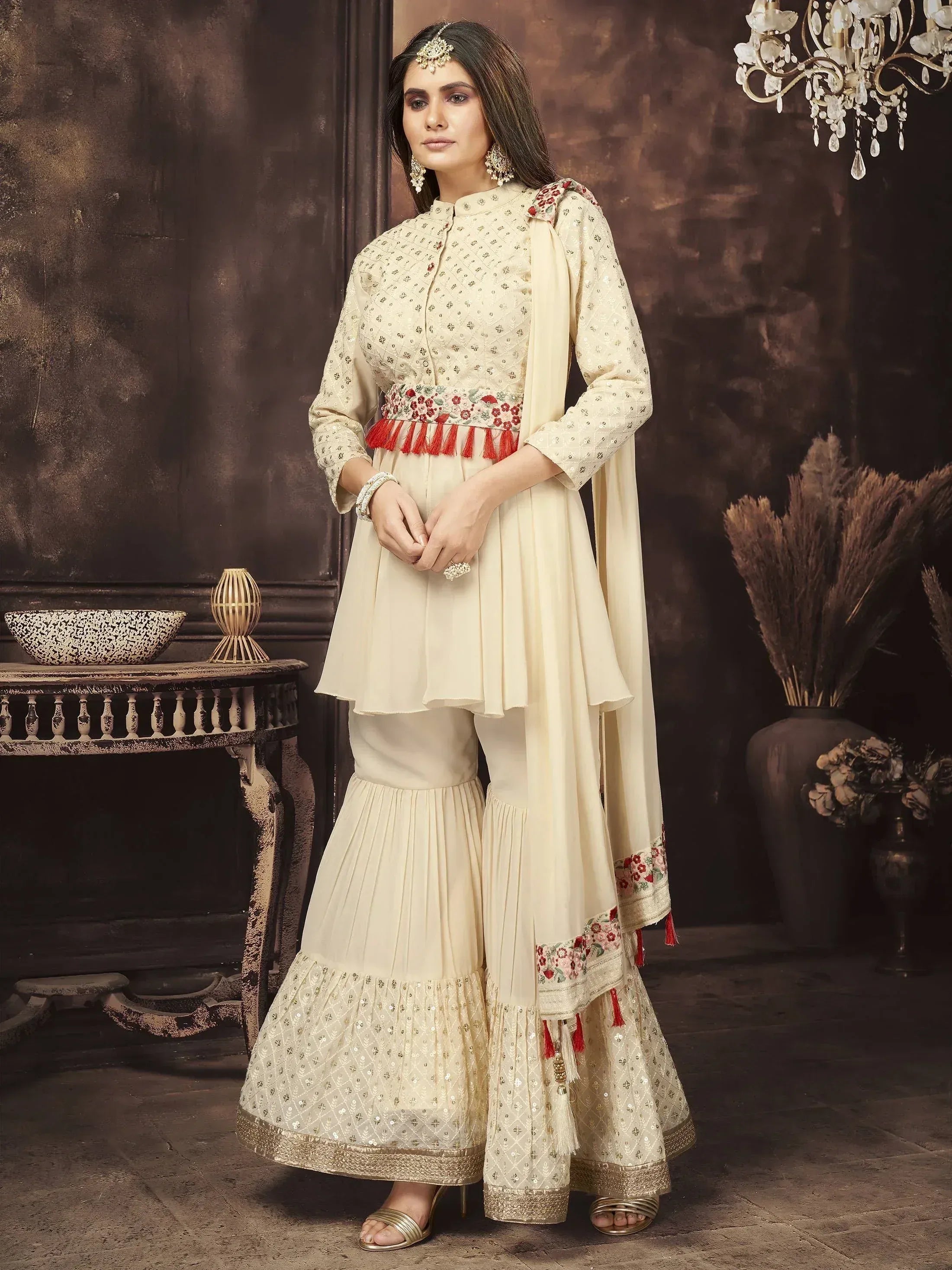 Woman wearing ivory embroidered Indian ethnic sharara suit with dupatta and jewelry indoors