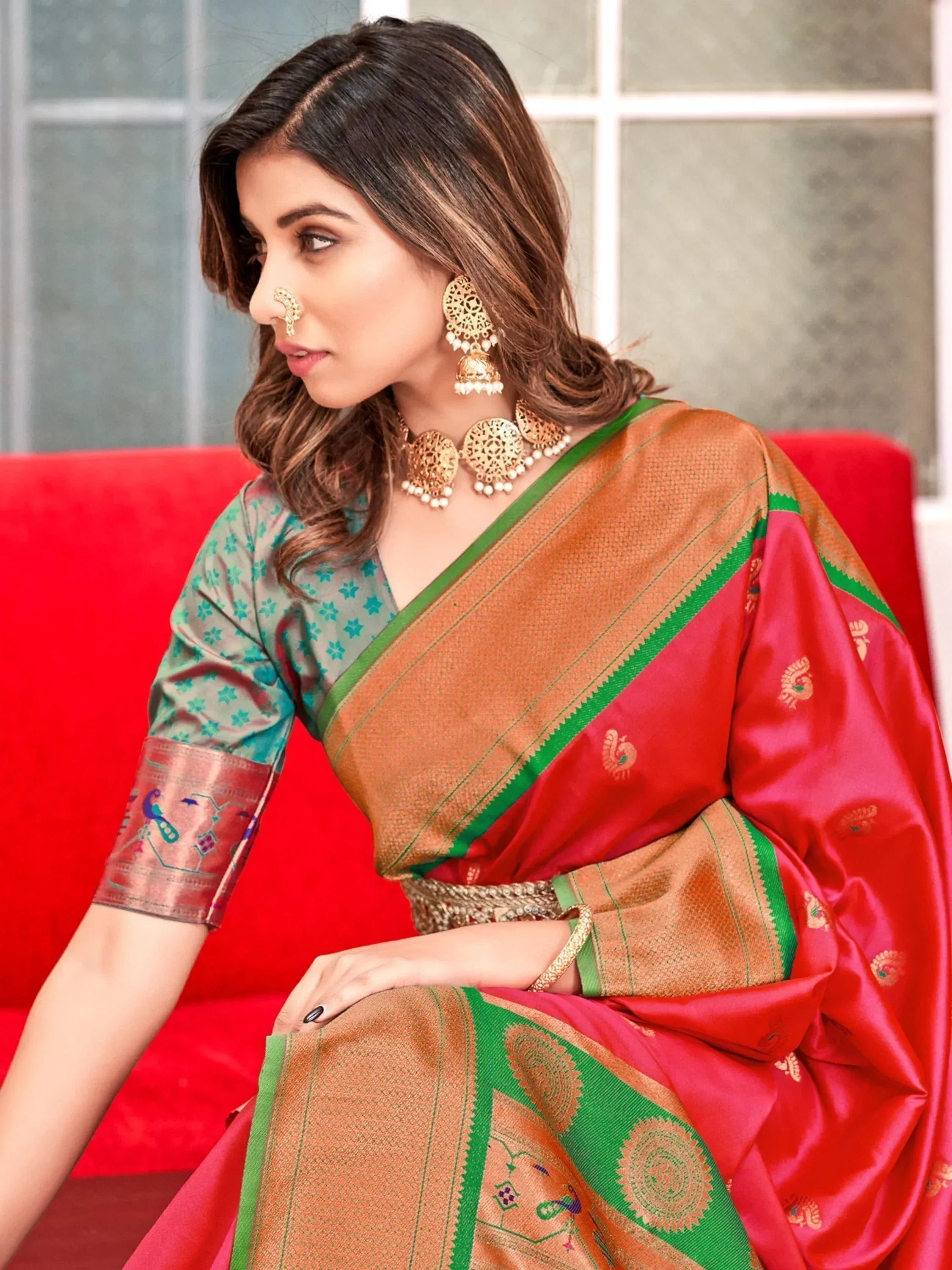 Woman in red and gold designer saree with green border, ethnic jewelry, sitting on red sofa