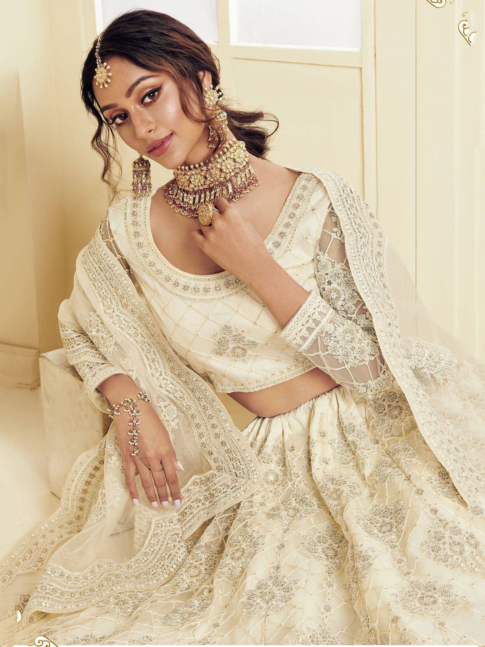 Elegant cream designer lehenga choli with intricate embroidery and matching gold jewelry for Indian weddings.