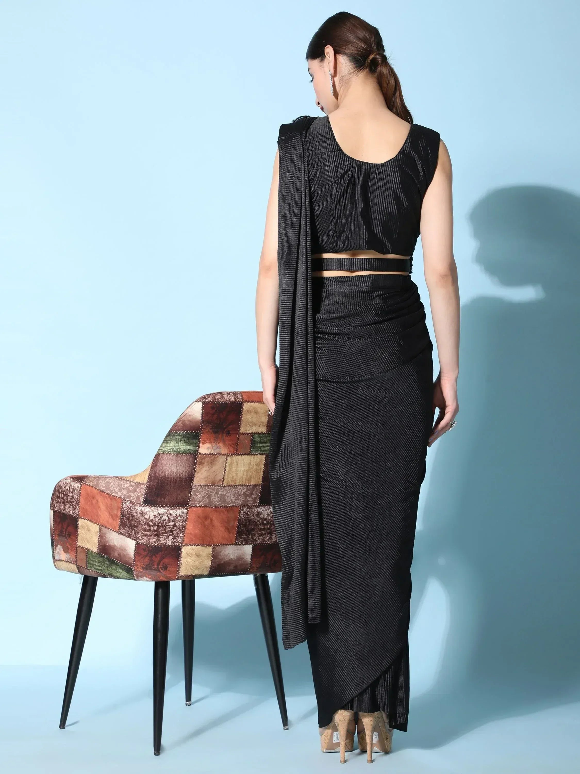 Black pleated ethnic gown with draped back, styled with high heels and patchwork chair