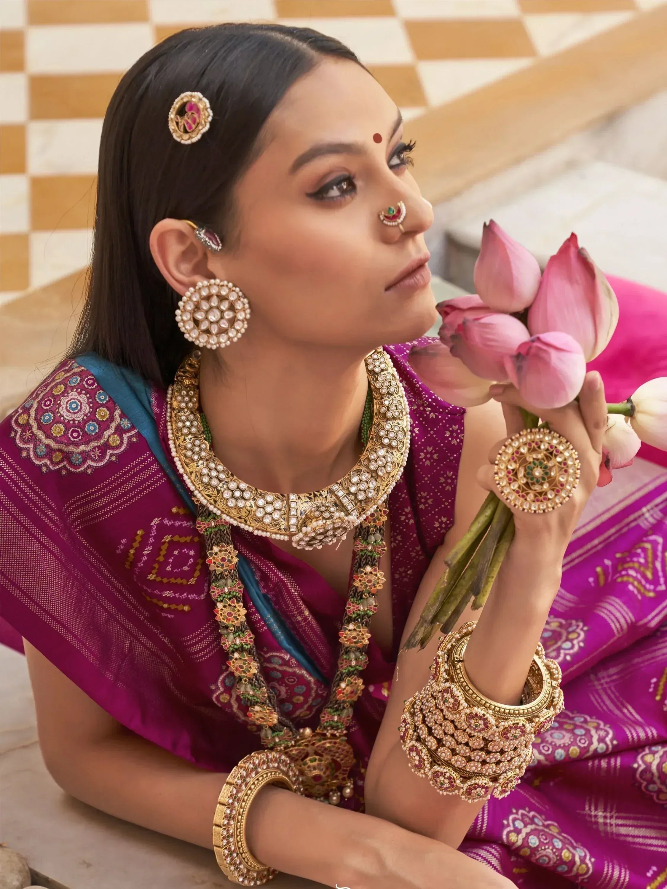 Woman in purple ethnic saree with gold jewelry holding pink lotus flowers, traditional Indian attire