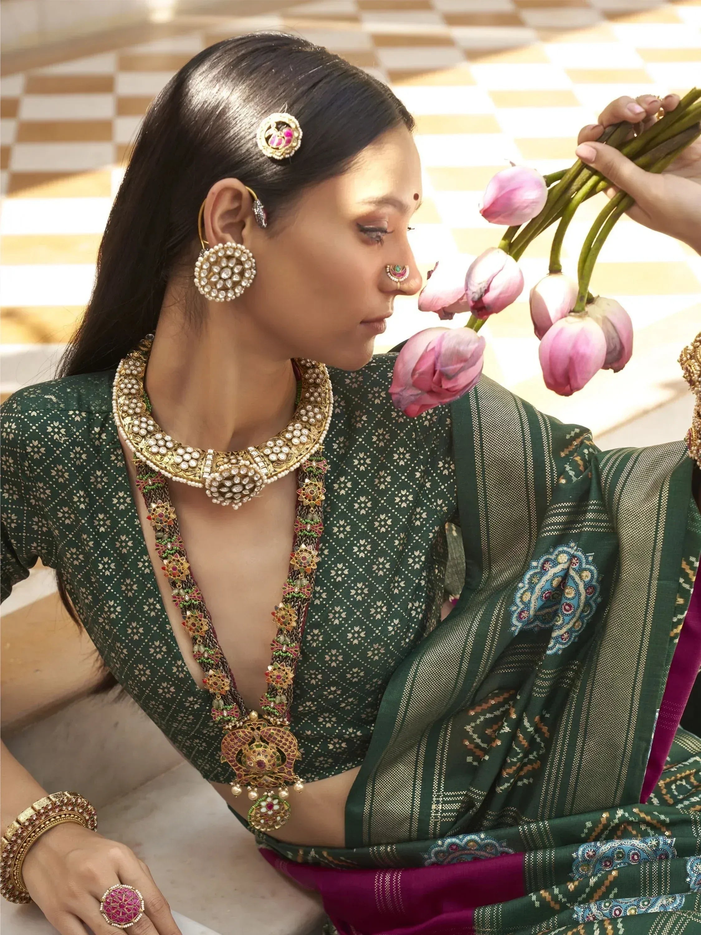 Elegant woman in green ethnic saree with intricate jewelry, holding pink lotus flowers, traditional Indian attire