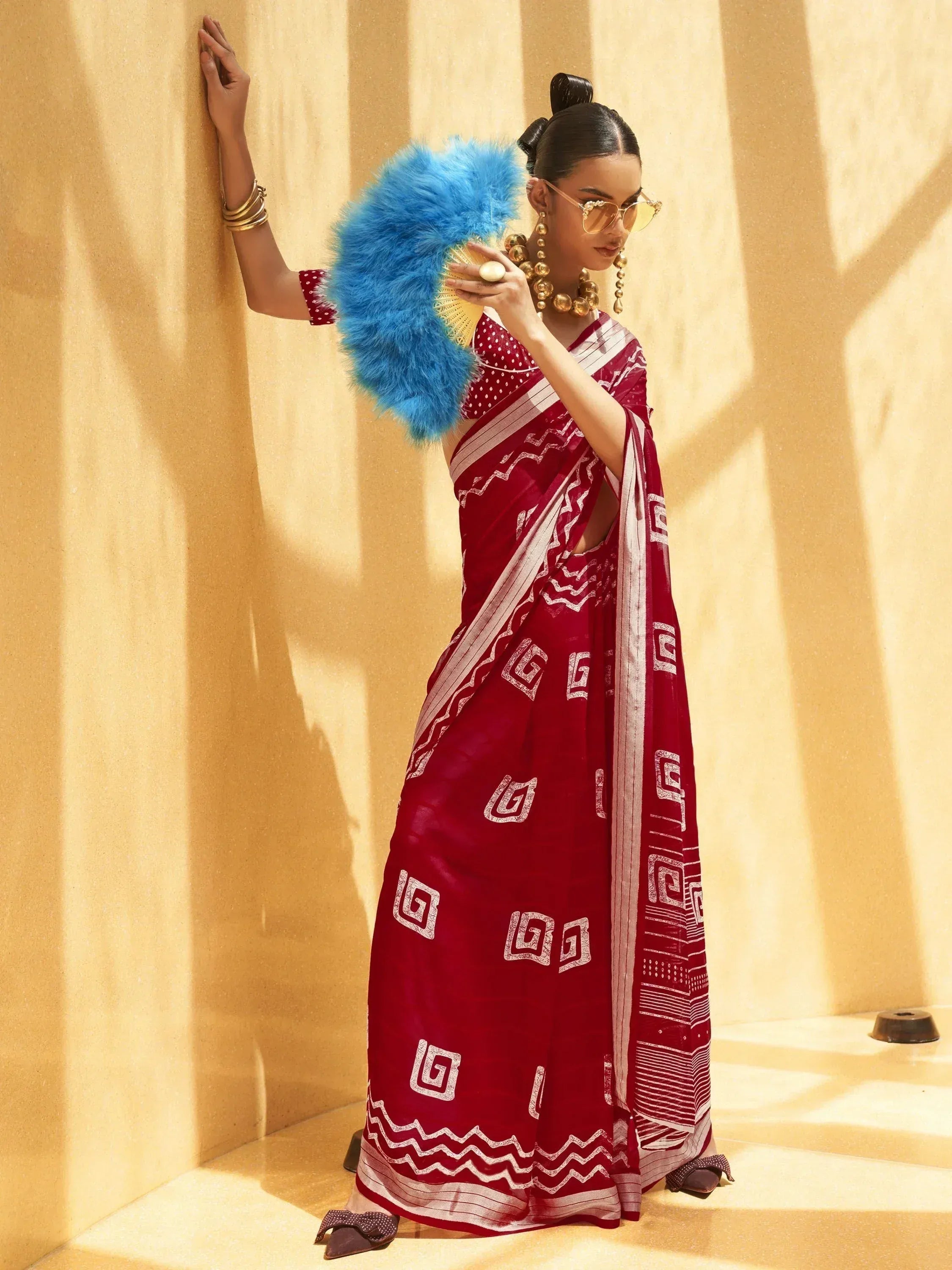 Woman in maroon printed saree with gold jewelry and blue feather fan, stylish ethnic wear