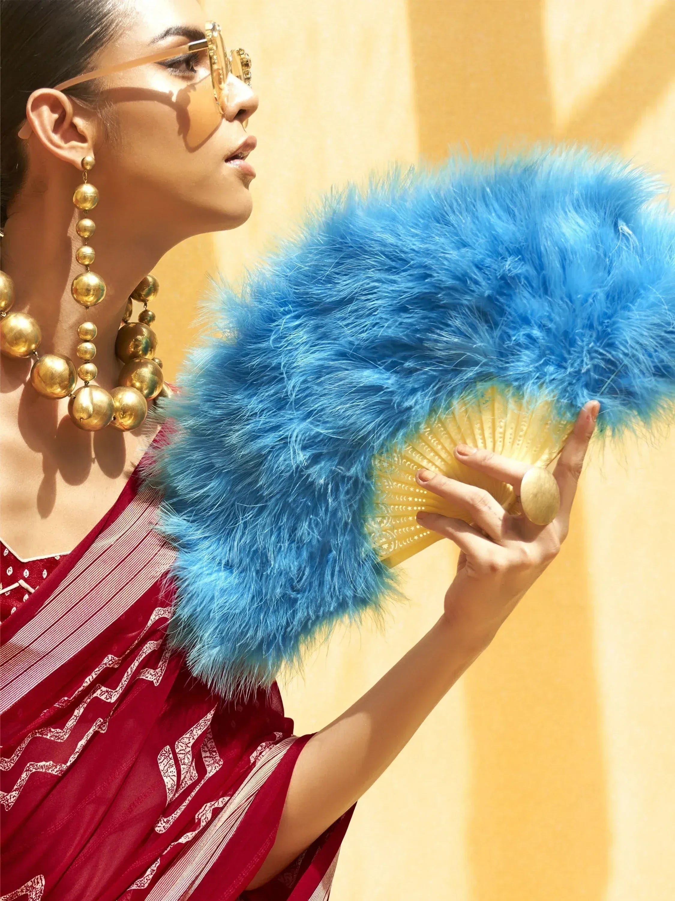 Woman in red ethnic saree with gold jewelry holding blue feather fan, stylish Indian fashion accessory