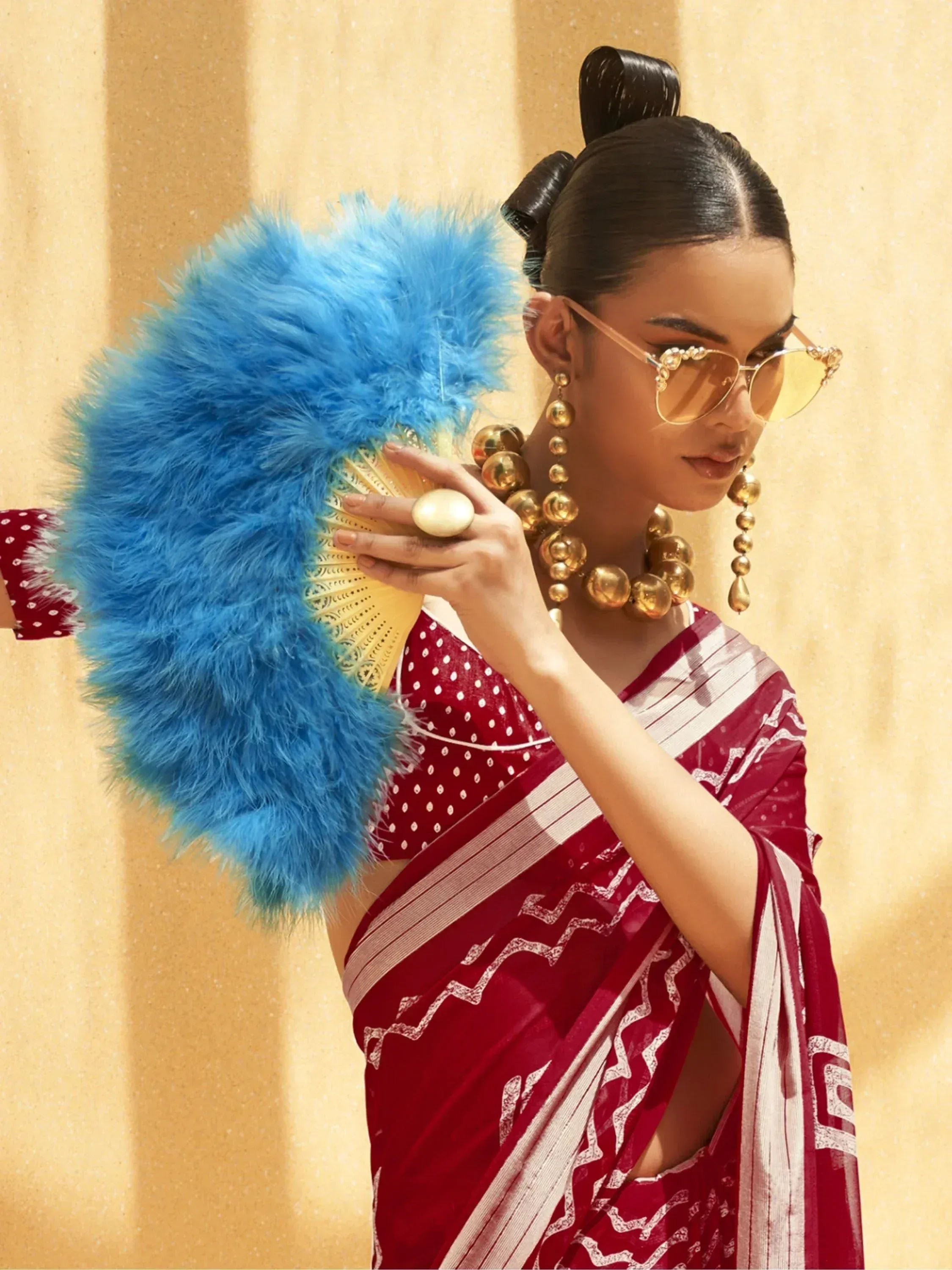 Woman in red printed saree with blue feather fan, gold jewelry, and sunglasses for ethnic fashion