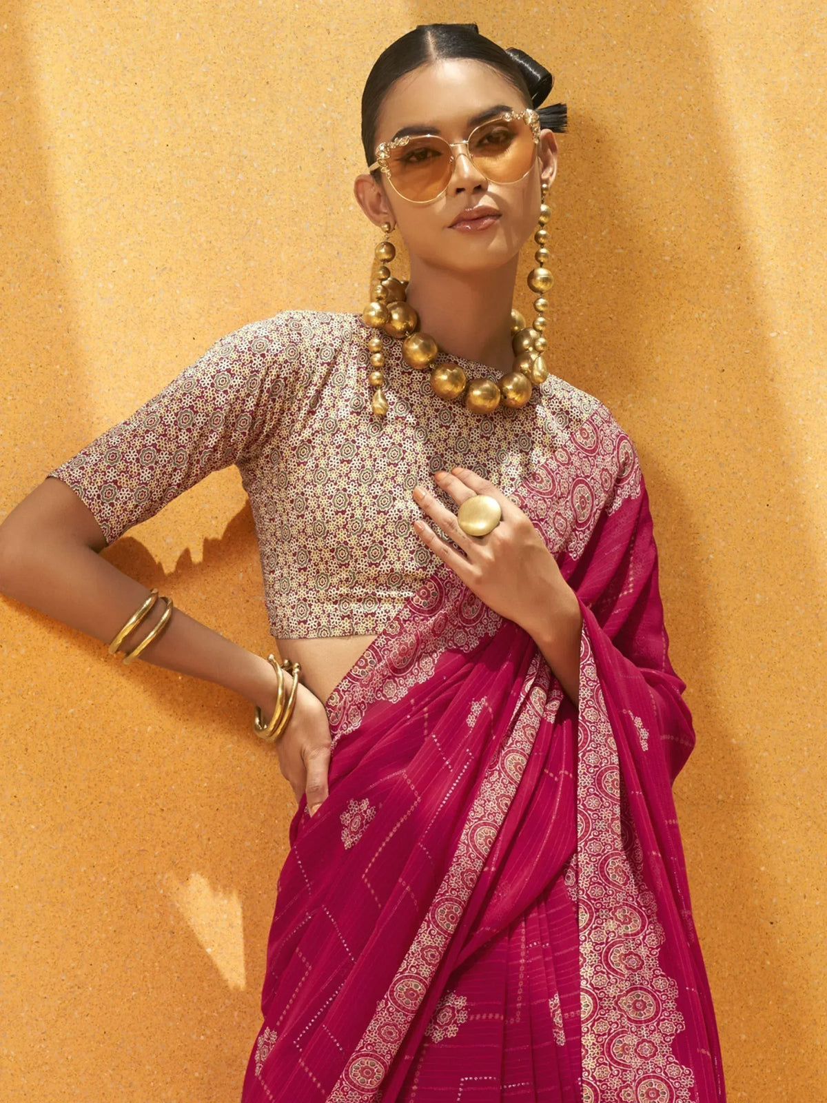 Woman in vibrant pink printed saree with gold jewelry and sunglasses, styled for ethnic wear