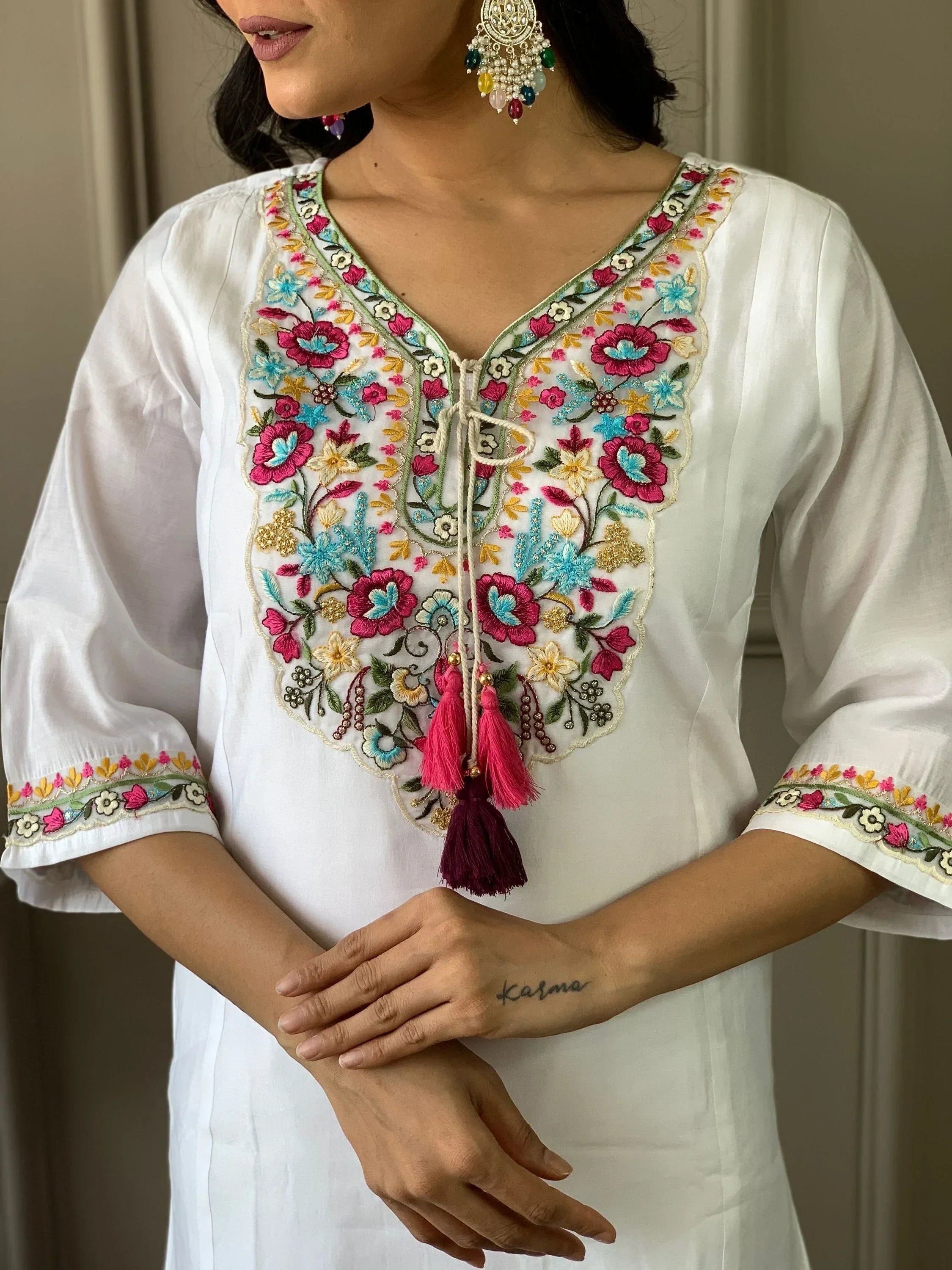 White kurti with colorful floral embroidery, mirror work, and tassels, worn with statement earrings