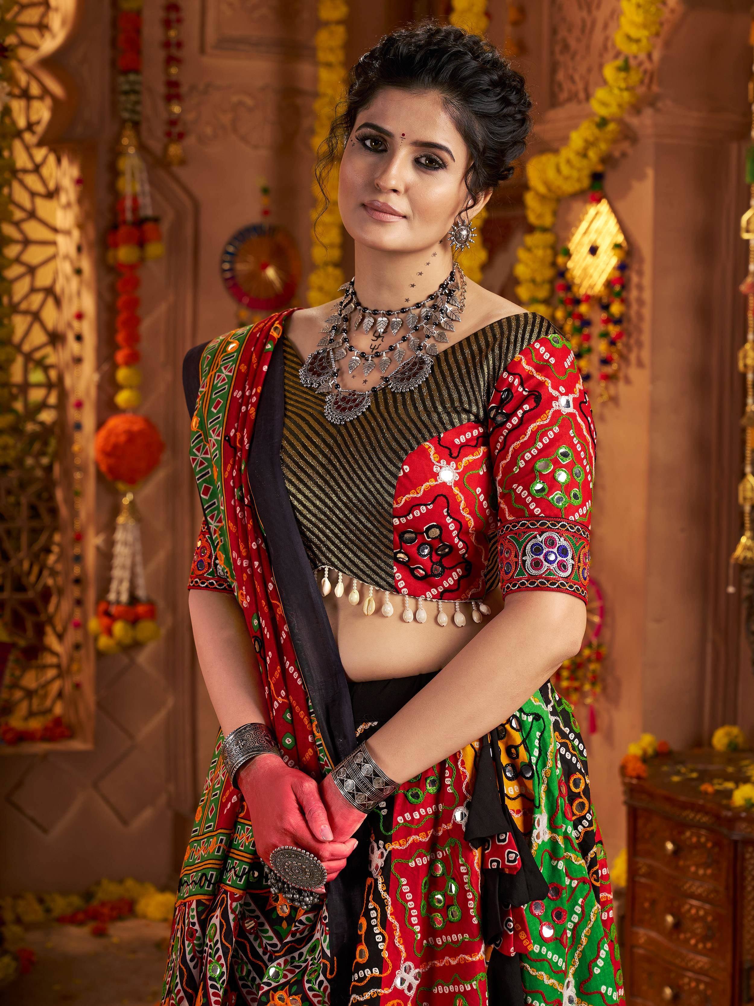Woman in vibrant red and green embroidered ethnic lehenga choli with mirror work, traditional jewelry, and festive floral backdrop