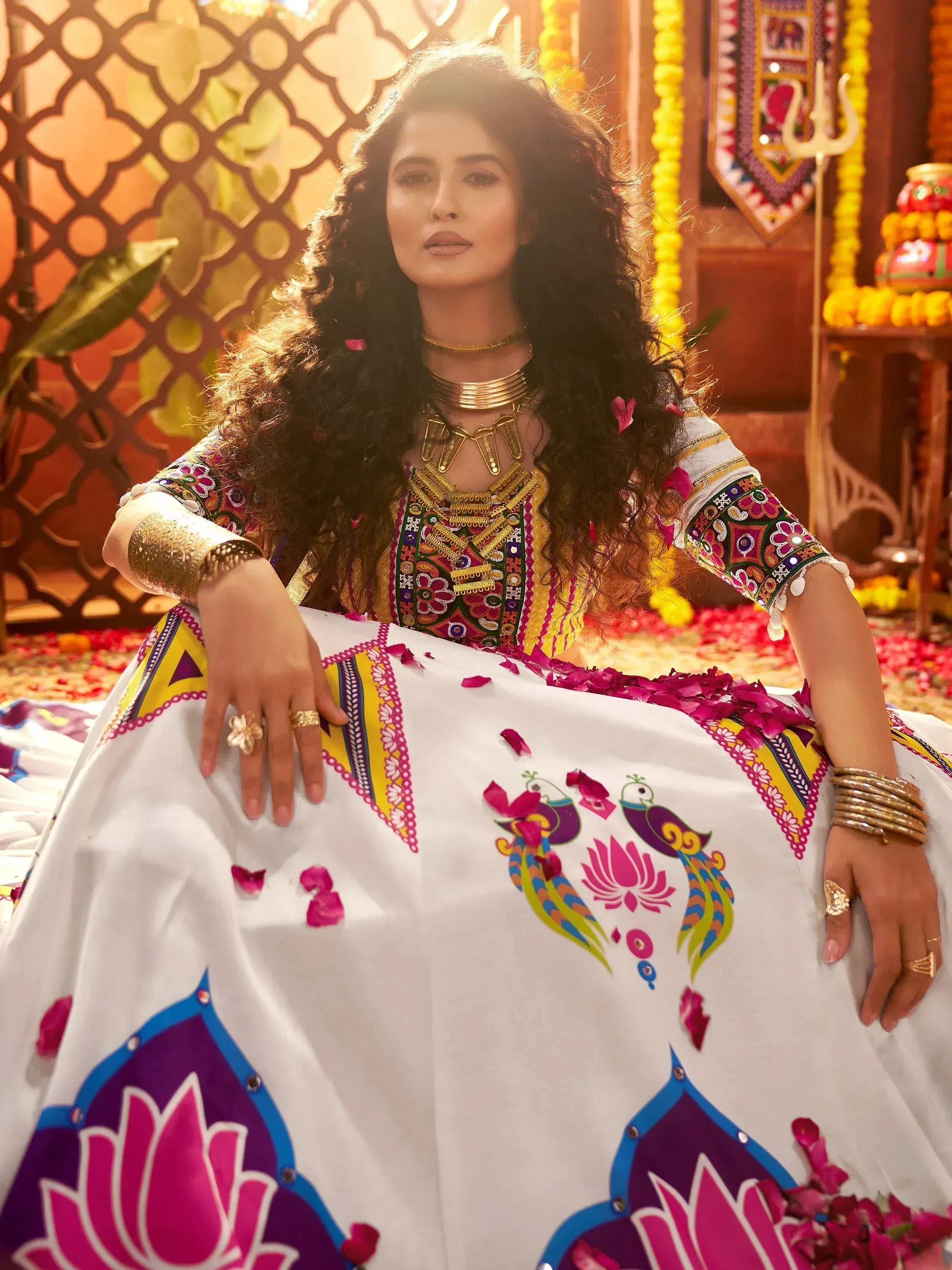 Woman in vibrant Indian ethnic gown with peacock and lotus print, adorned with gold jewelry and floral petals