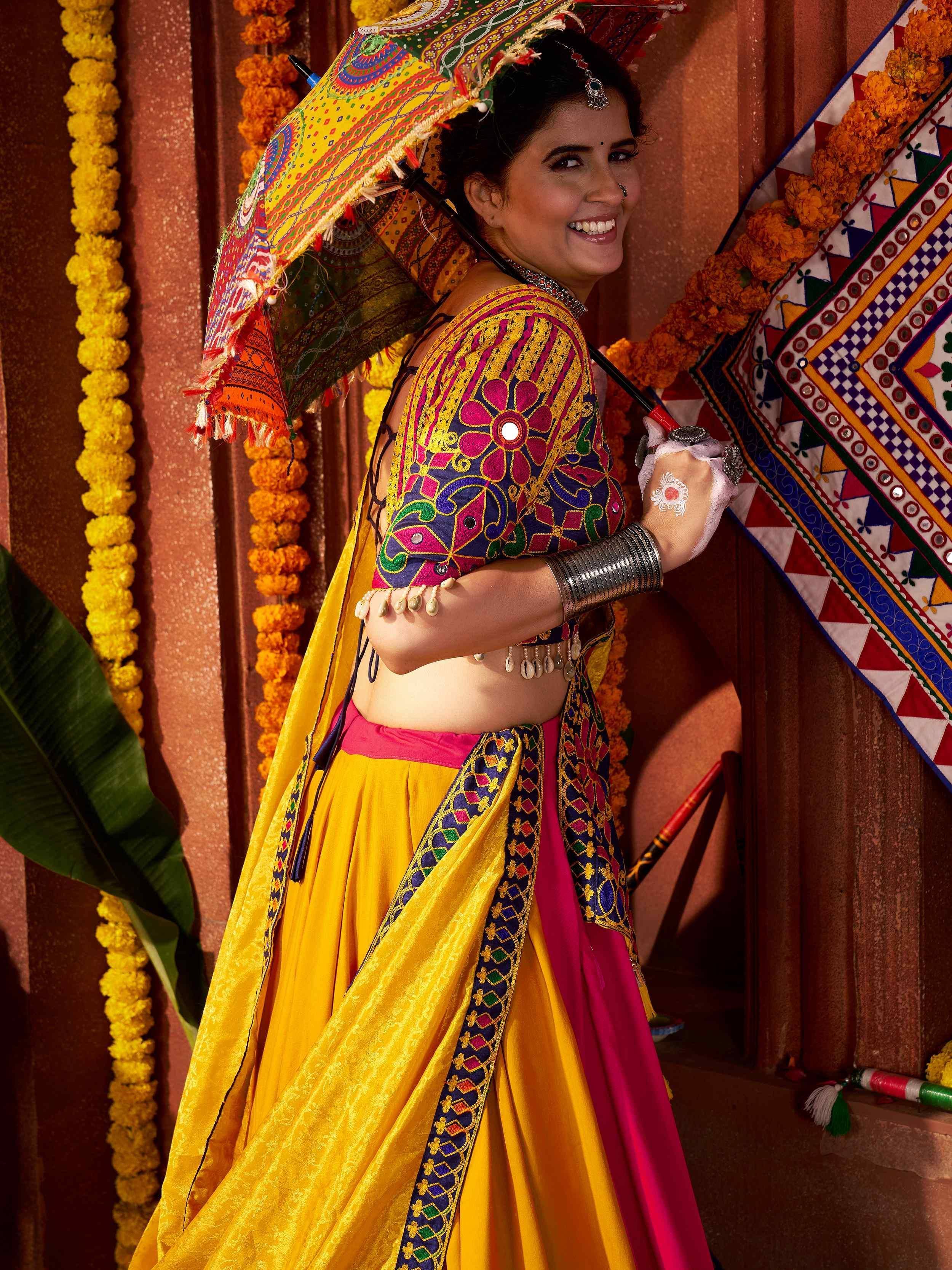 Woman in vibrant yellow and pink embroidered lehenga choli with colorful umbrella, festive Indian ethnic wear
