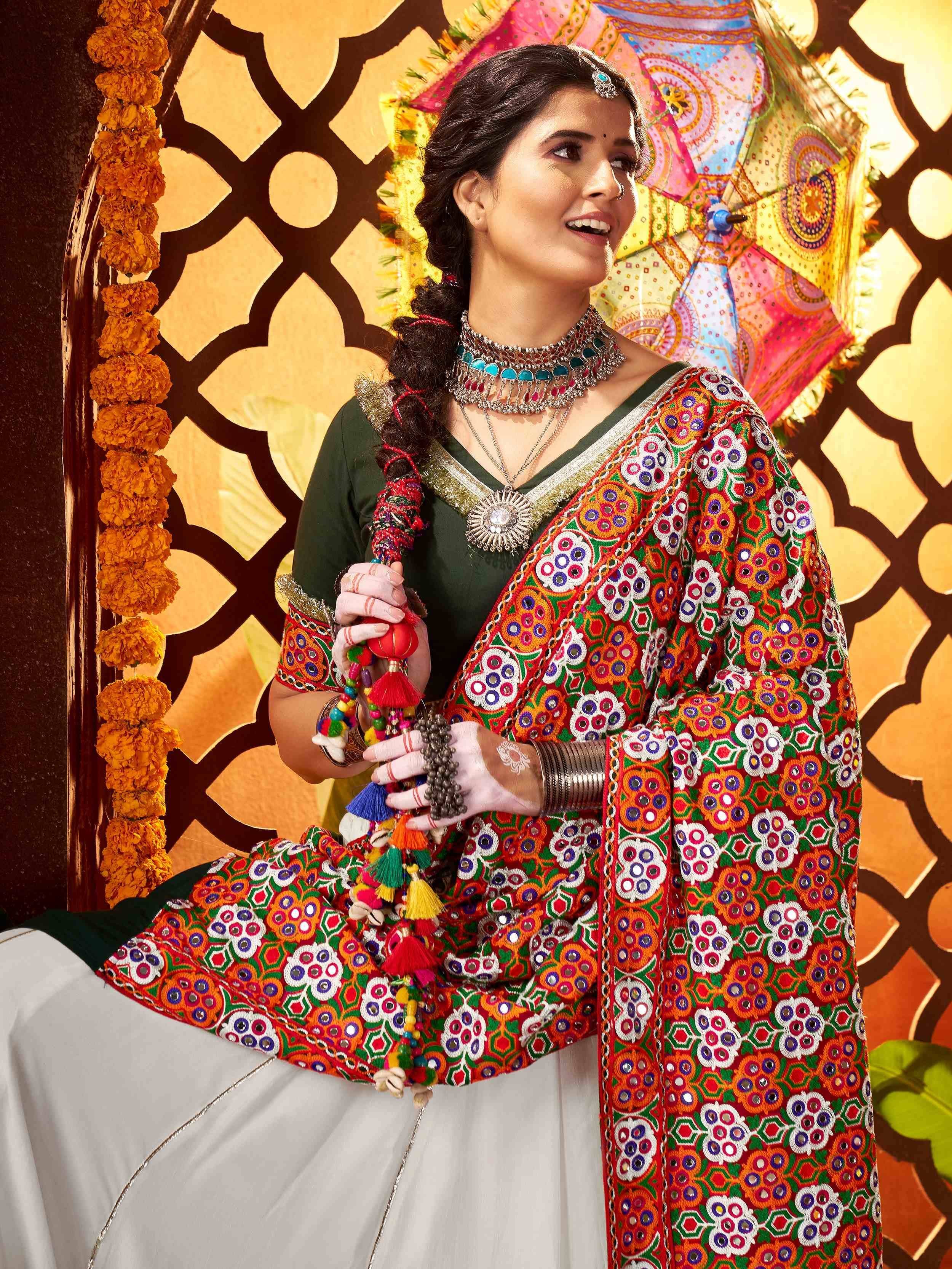 Woman in vibrant Indian ethnic wear with mirror work, floral patterns, and traditional jewelry, holding colorful tassels.
