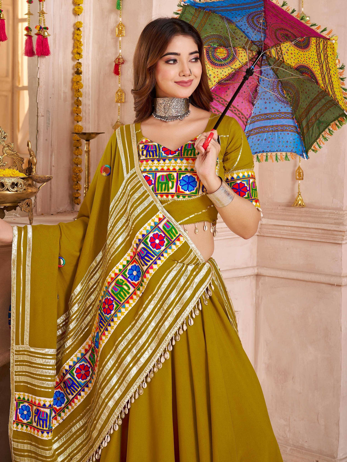 Woman in olive green embroidered lehenga choli with colorful mirror work, holding a traditional umbrella.