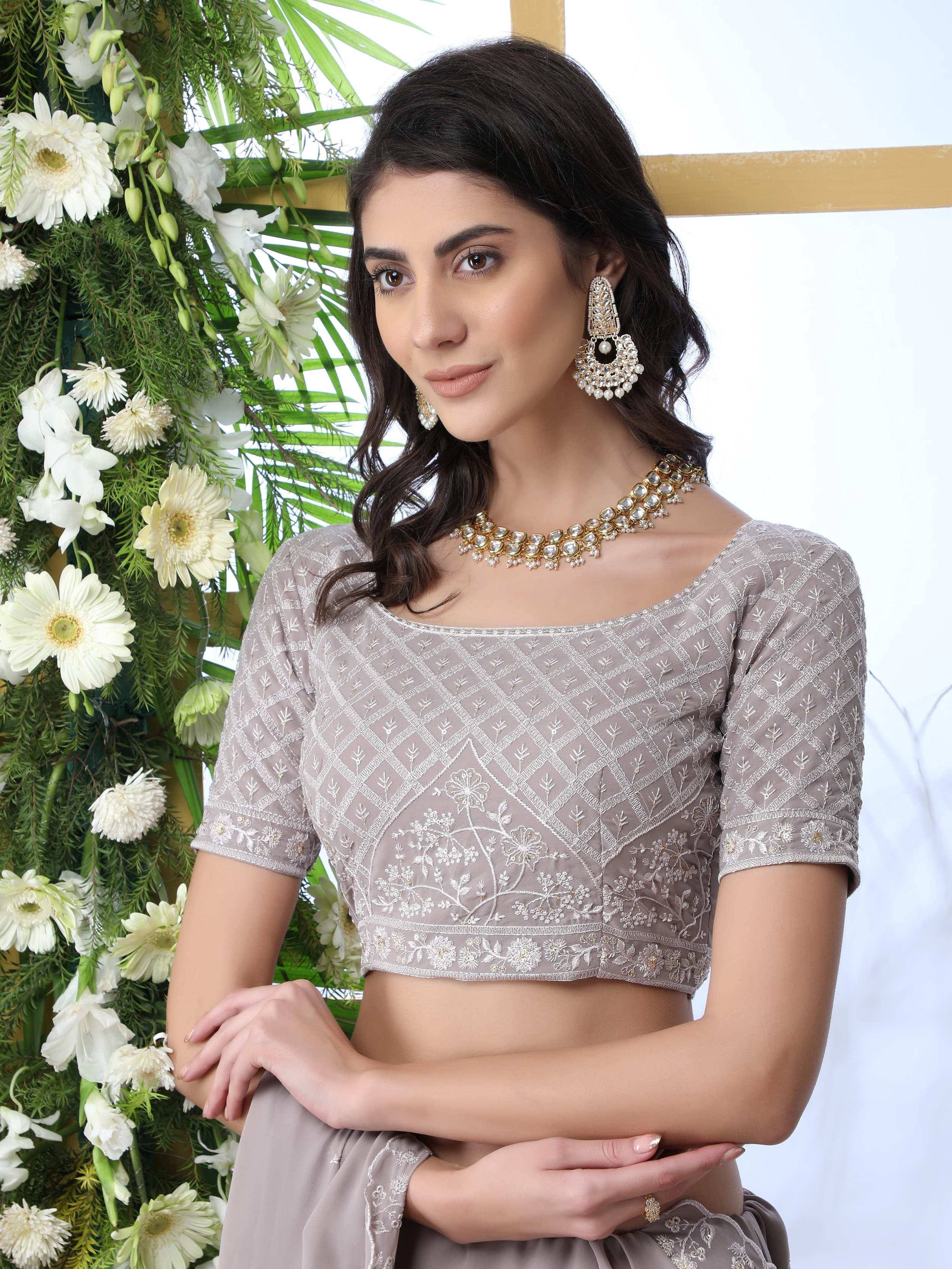 Elegant grey embroidered lehenga choli blouse with floral detailing, paired with gold jewelry and white floral backdrop.