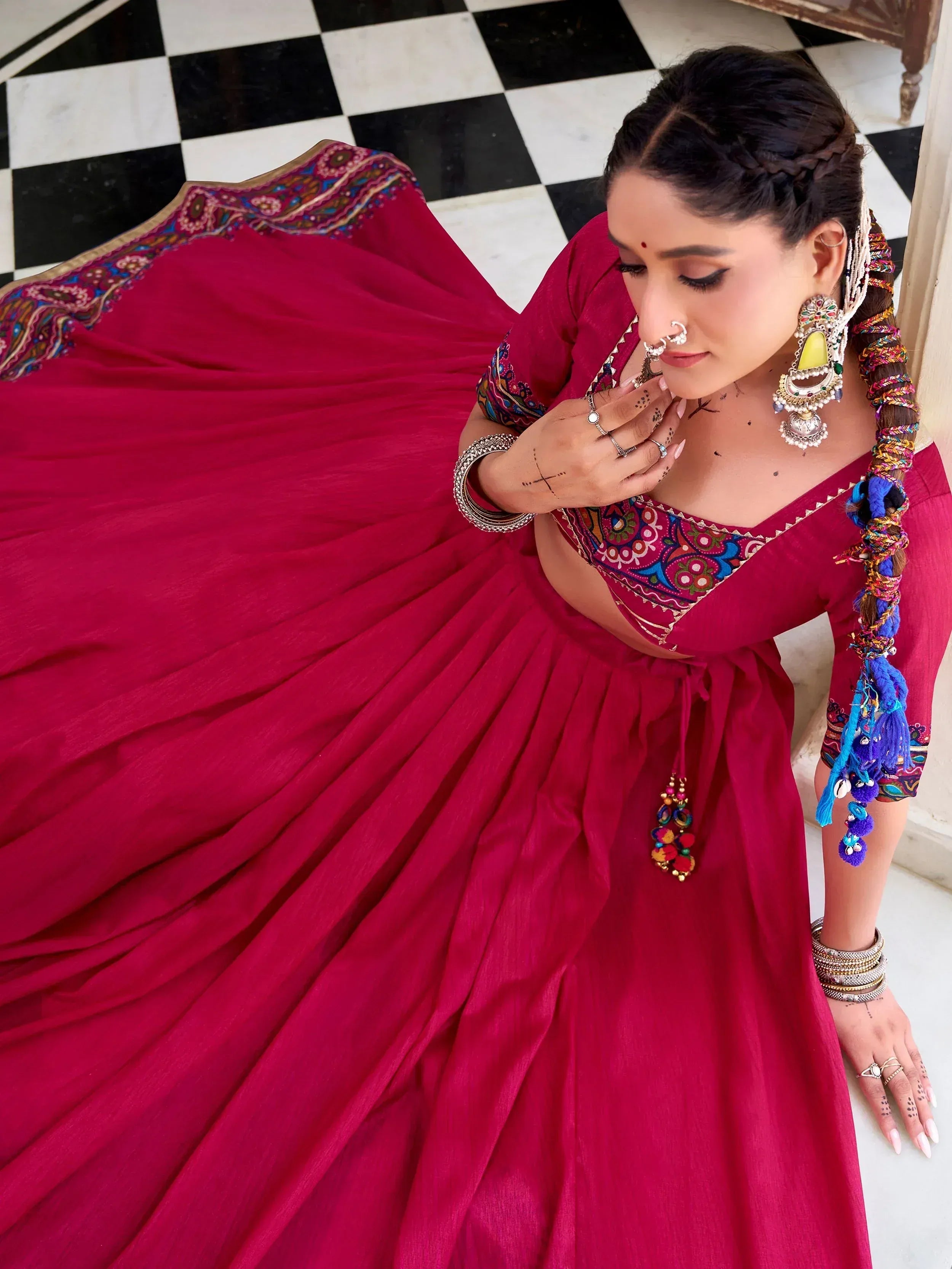 Woman in vibrant red ethnic lehenga choli with colorful embroidery, traditional jewelry, and braided hair on checkered floor