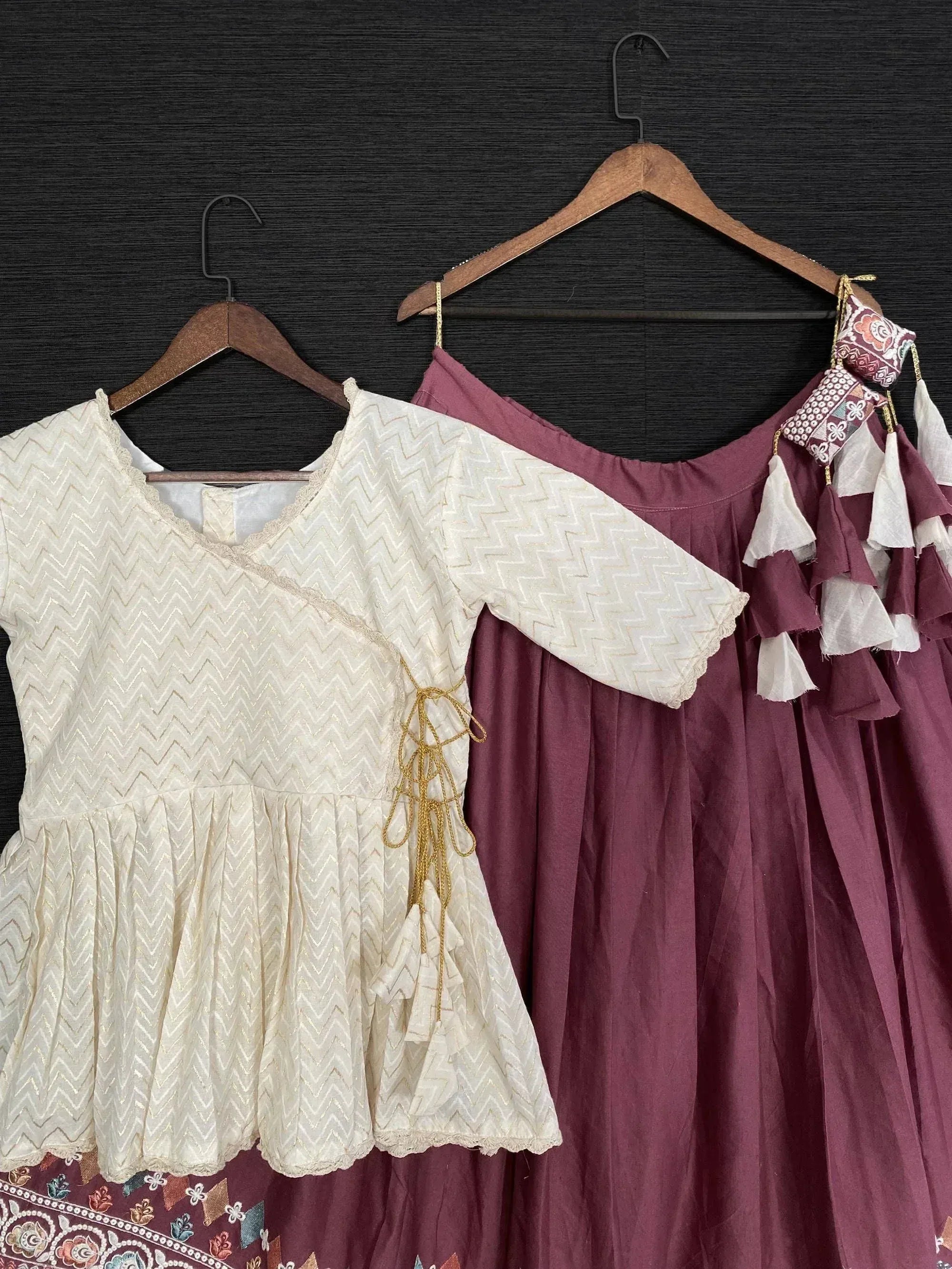 Cream zigzag print top with gold lace-up detail and maroon ethnic gown with tassel trim, hanging on wooden hangers
