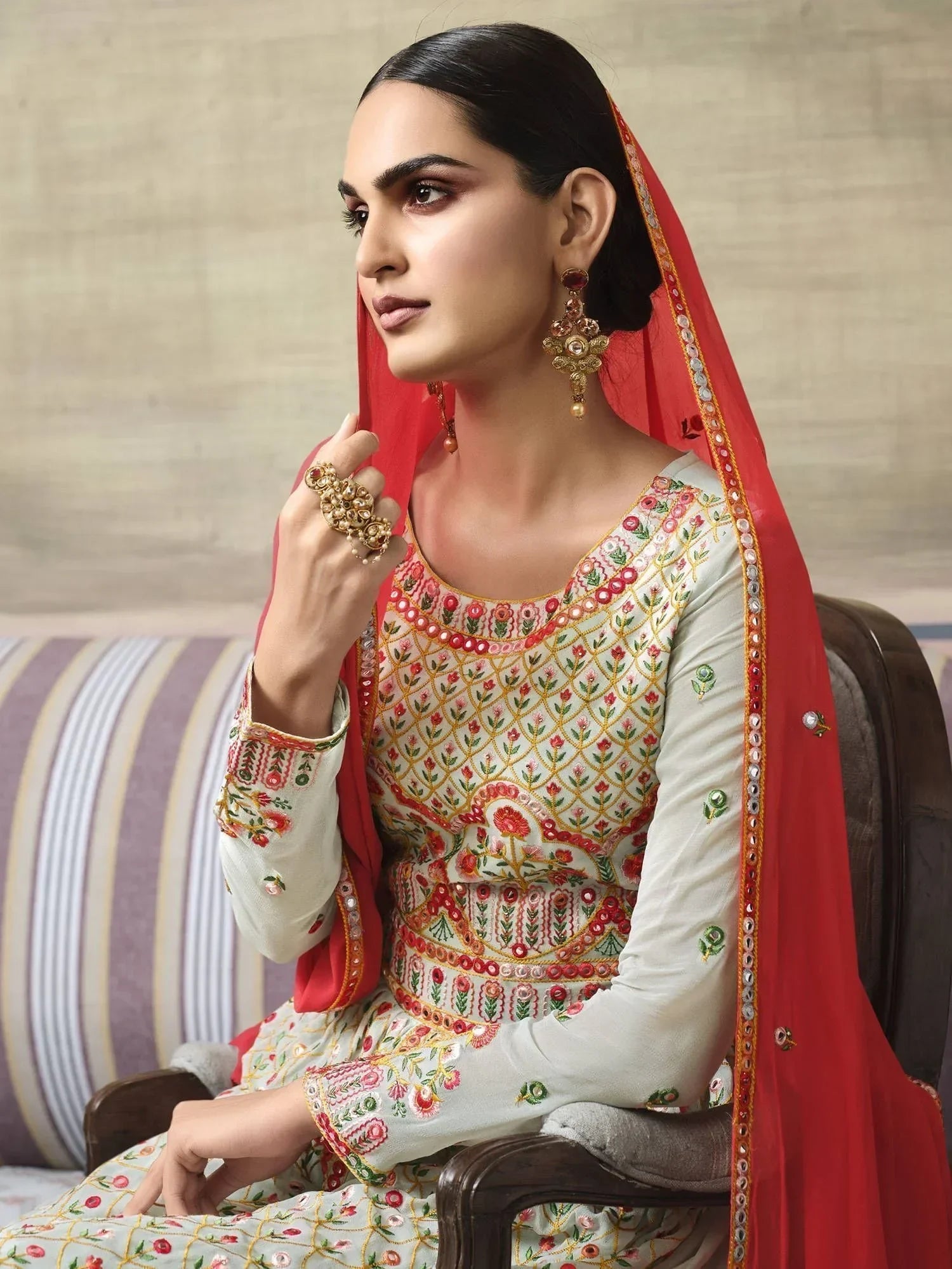 Woman in embroidered Anarkali suit with red dupatta, wearing statement jewelry, sitting indoors