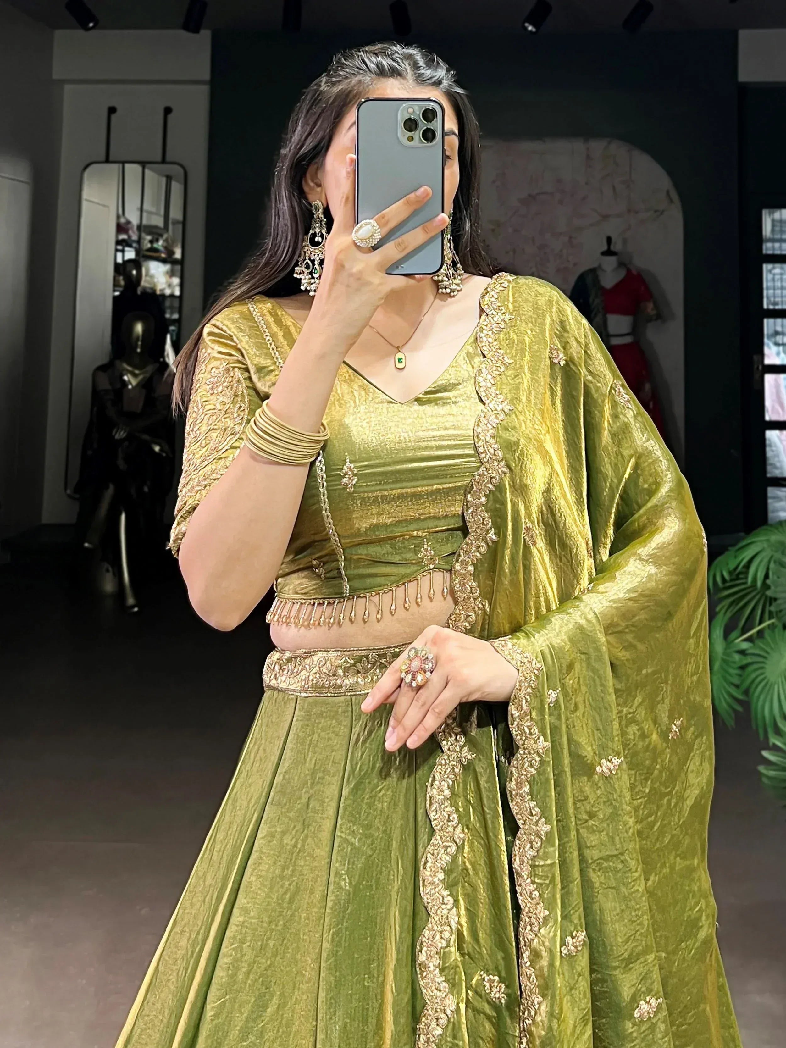 Woman in olive green velvet lehenga choli with gold embroidery, mirror selfie in boutique