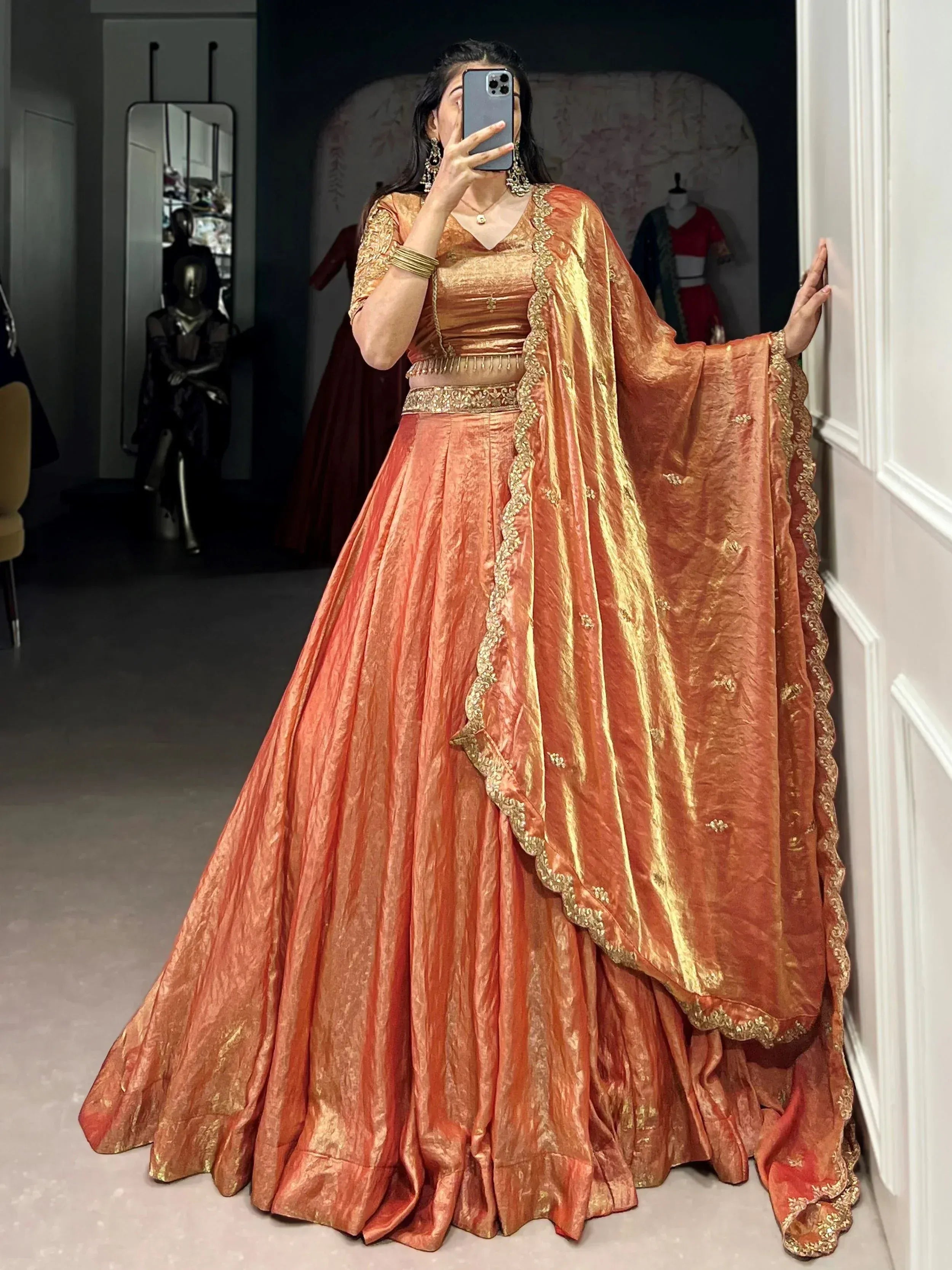 Woman in orange-gold designer lehenga choli with gold embroidery, taking mirror selfie in boutique