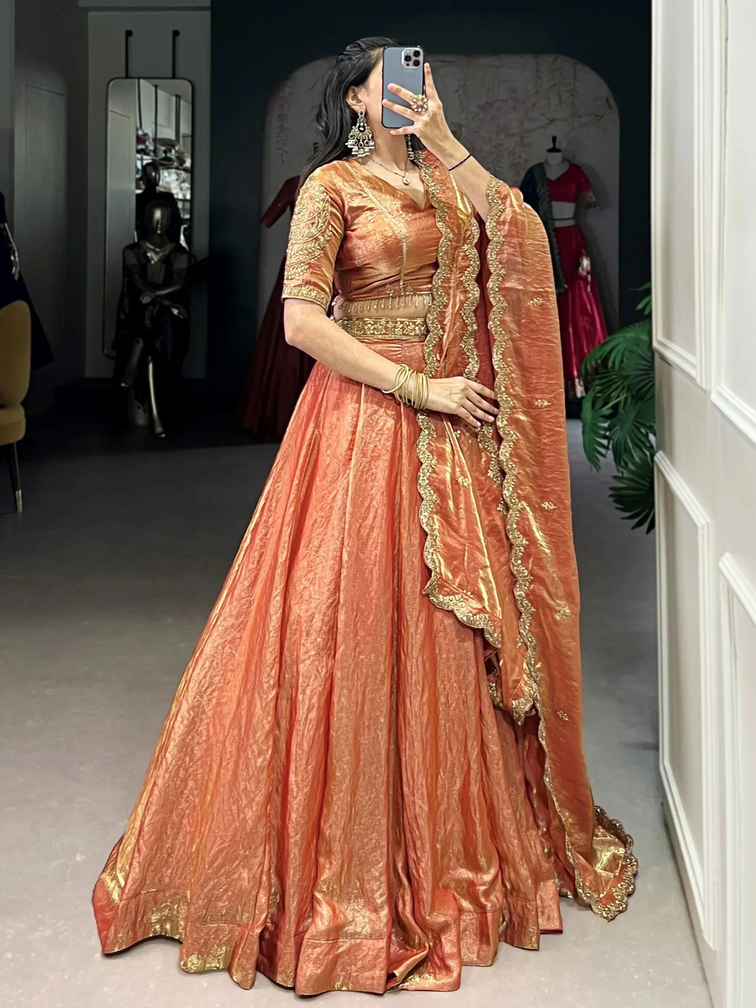 Woman in orange designer lehenga choli with gold embroidery, taking mirror selfie in boutique