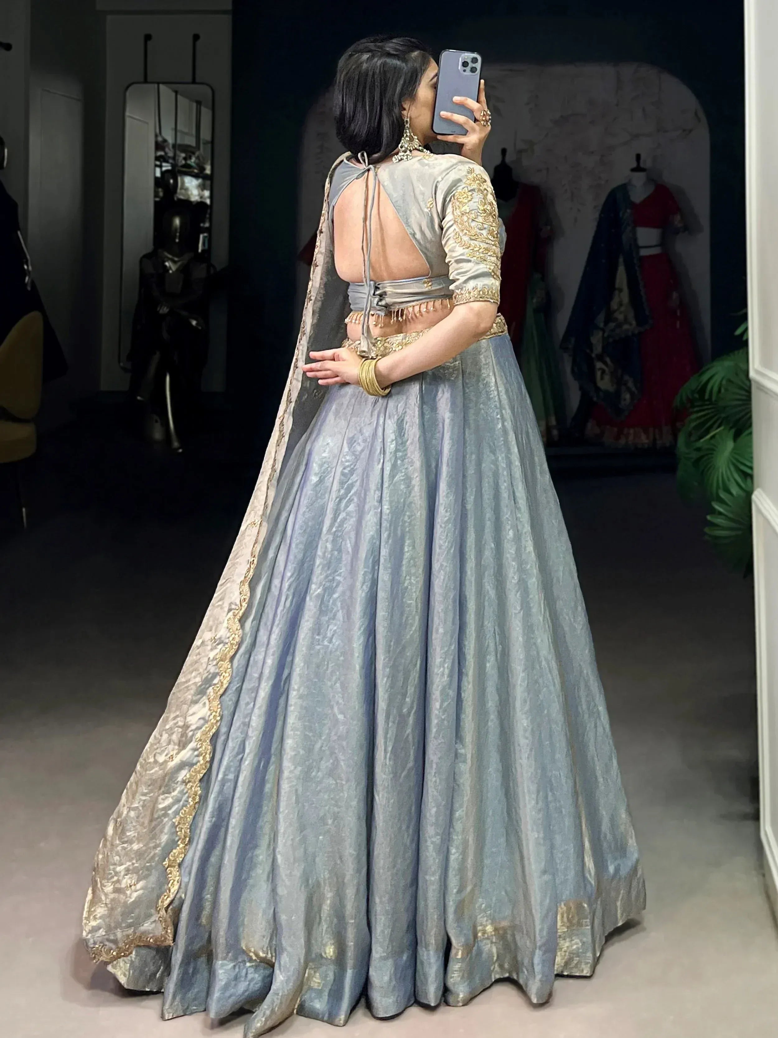 Woman in grey designer lehenga choli with gold embroidery, backless blouse, and dupatta, taking mirror selfie