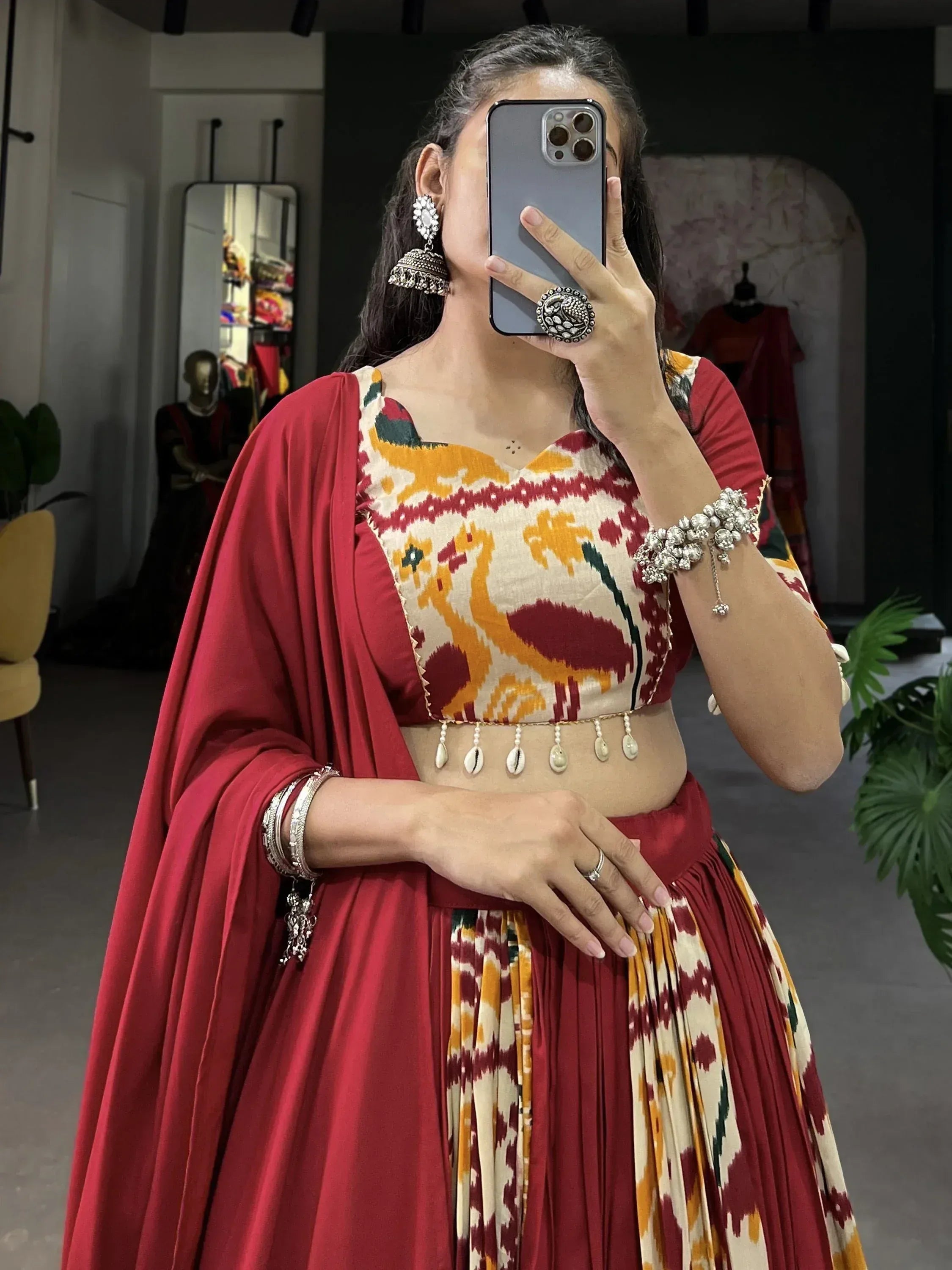 Woman in red and ikat print lehenga choli with statement jewelry, taking mirror selfie in boutique