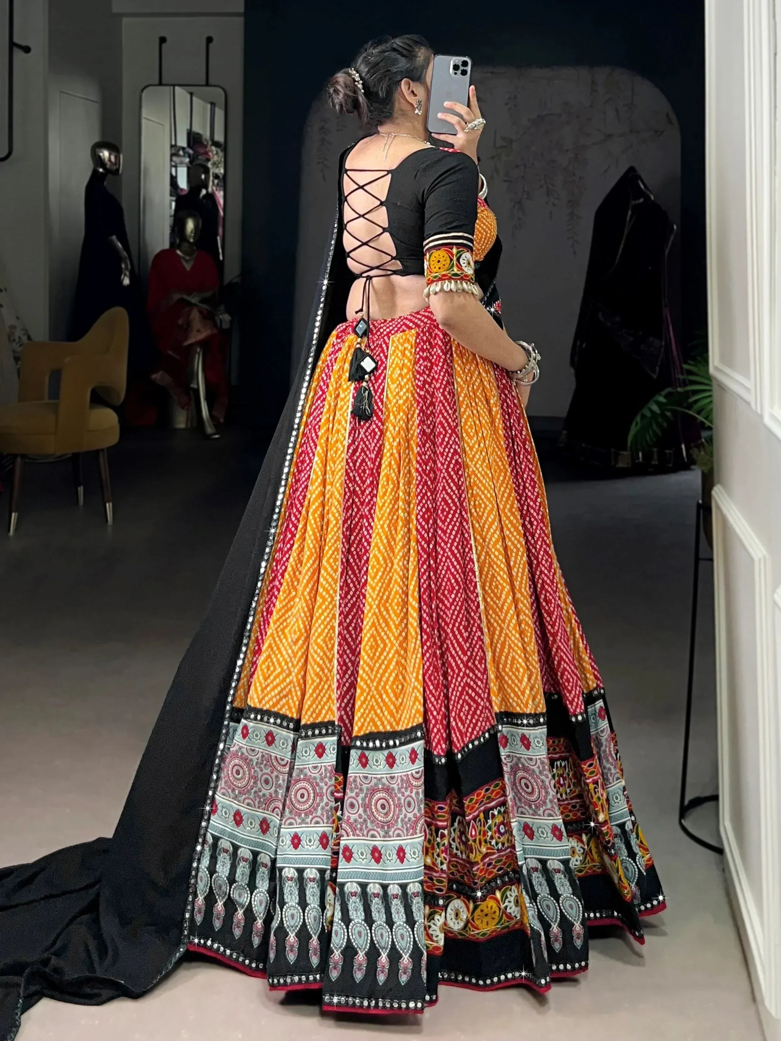 Woman in vibrant red and orange printed lehenga choli with black lace-up back and dupatta, taking mirror selfie
