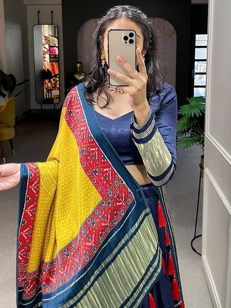 Woman in vibrant yellow and blue ethnic saree with red border, gold blouse, and tassels, taking mirror selfie.