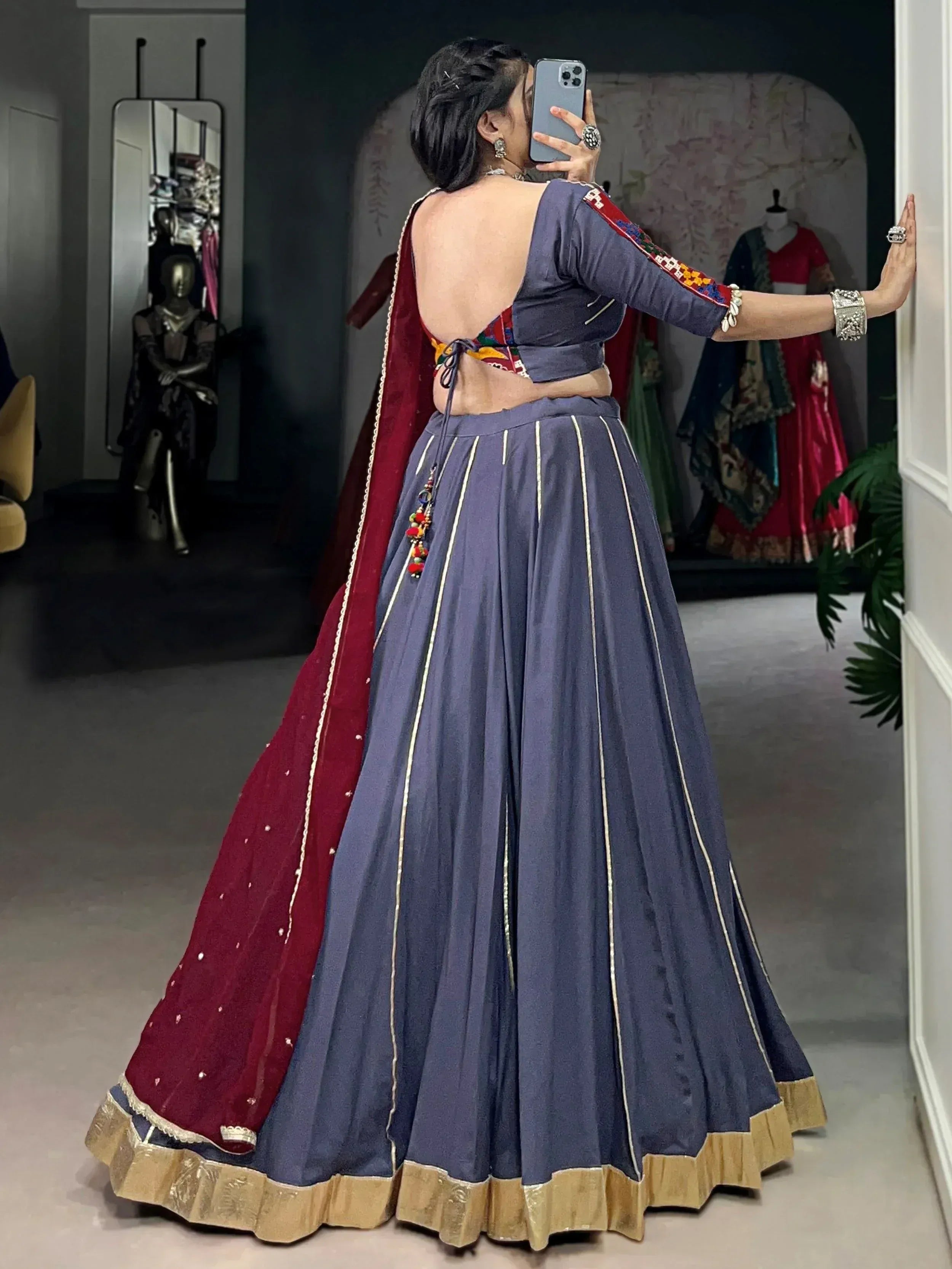 Woman in blue lehenga choli with open back, red dupatta, and gold trim, taking mirror selfie