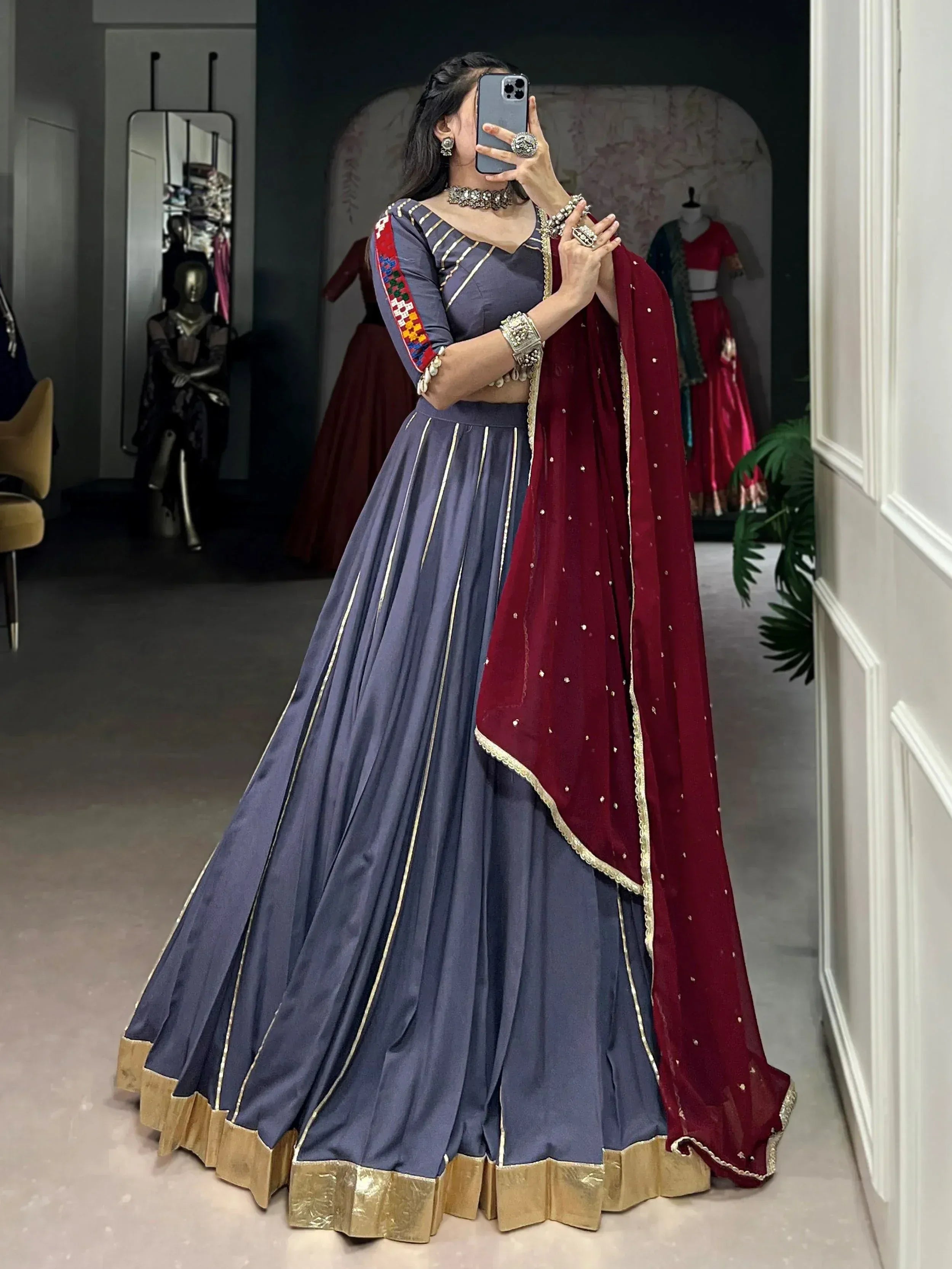 Woman in navy blue lehenga choli with gold trim and maroon dupatta, taking mirror selfie in boutique