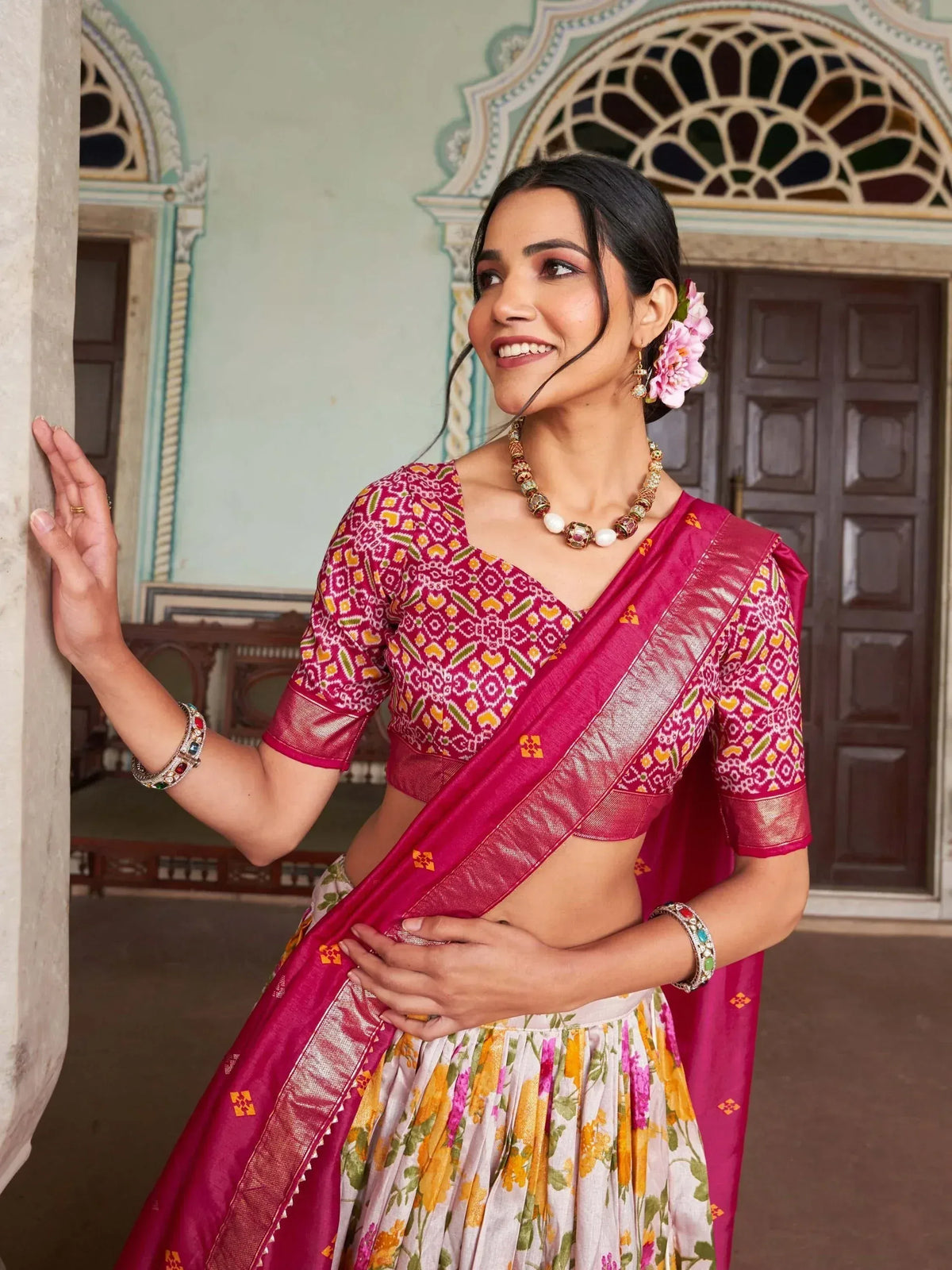 Woman in vibrant pink ethnic saree with floral lehenga, traditional jewelry, and temple backdrop