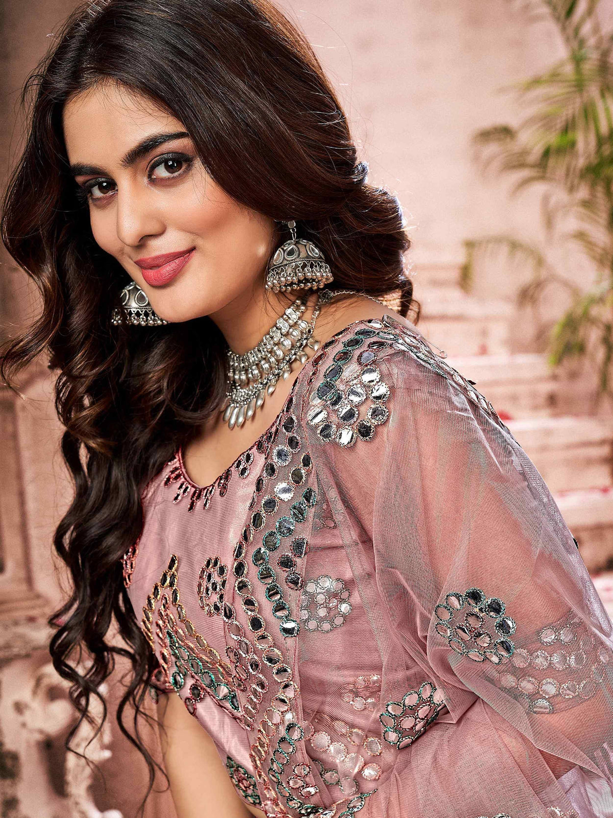Woman in pink ethnic gown with mirror work, paired with traditional jewelry for festive occasions.