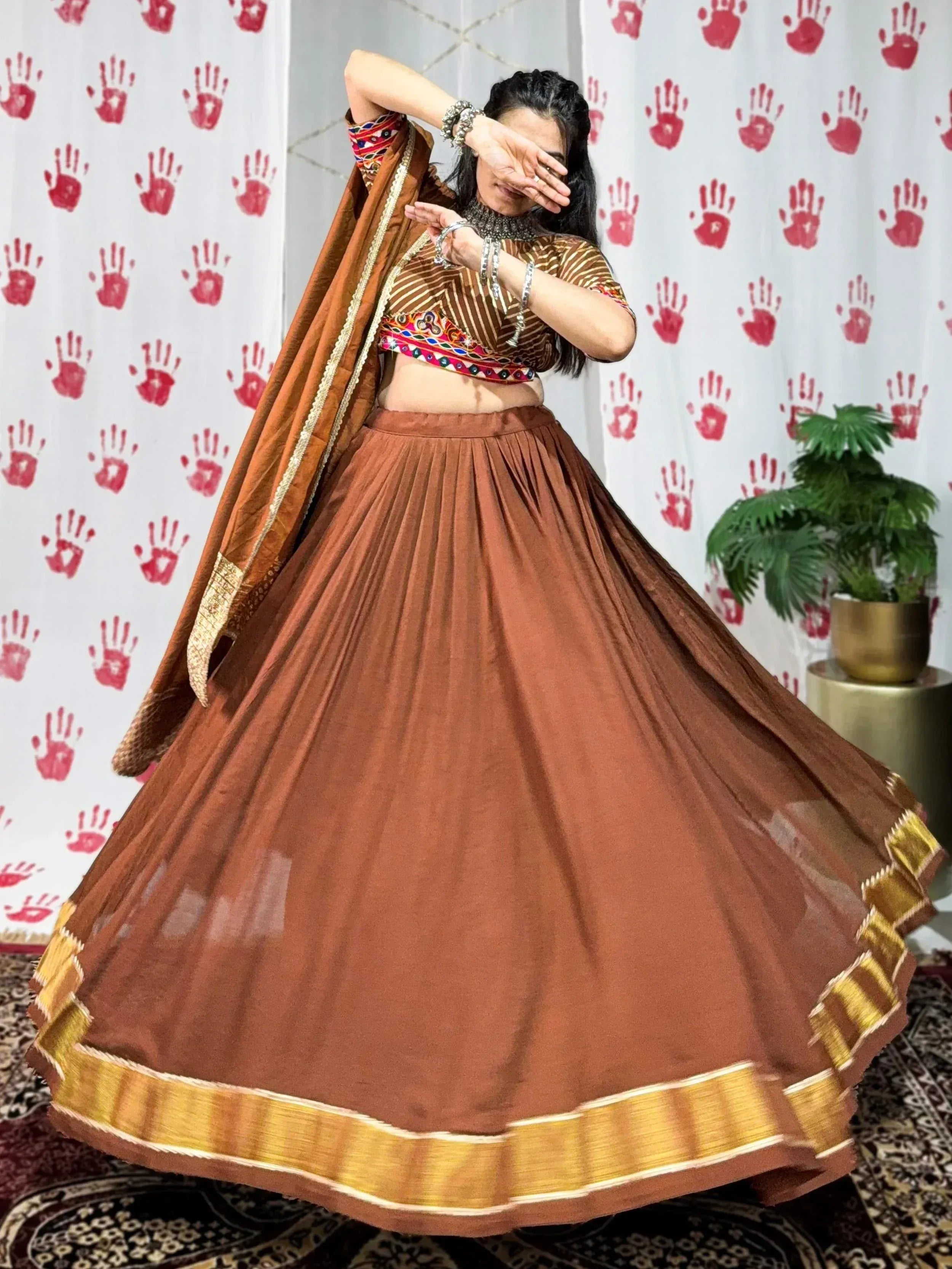 Woman in brown lehenga choli with gold border, twirling against red handprint backdrop