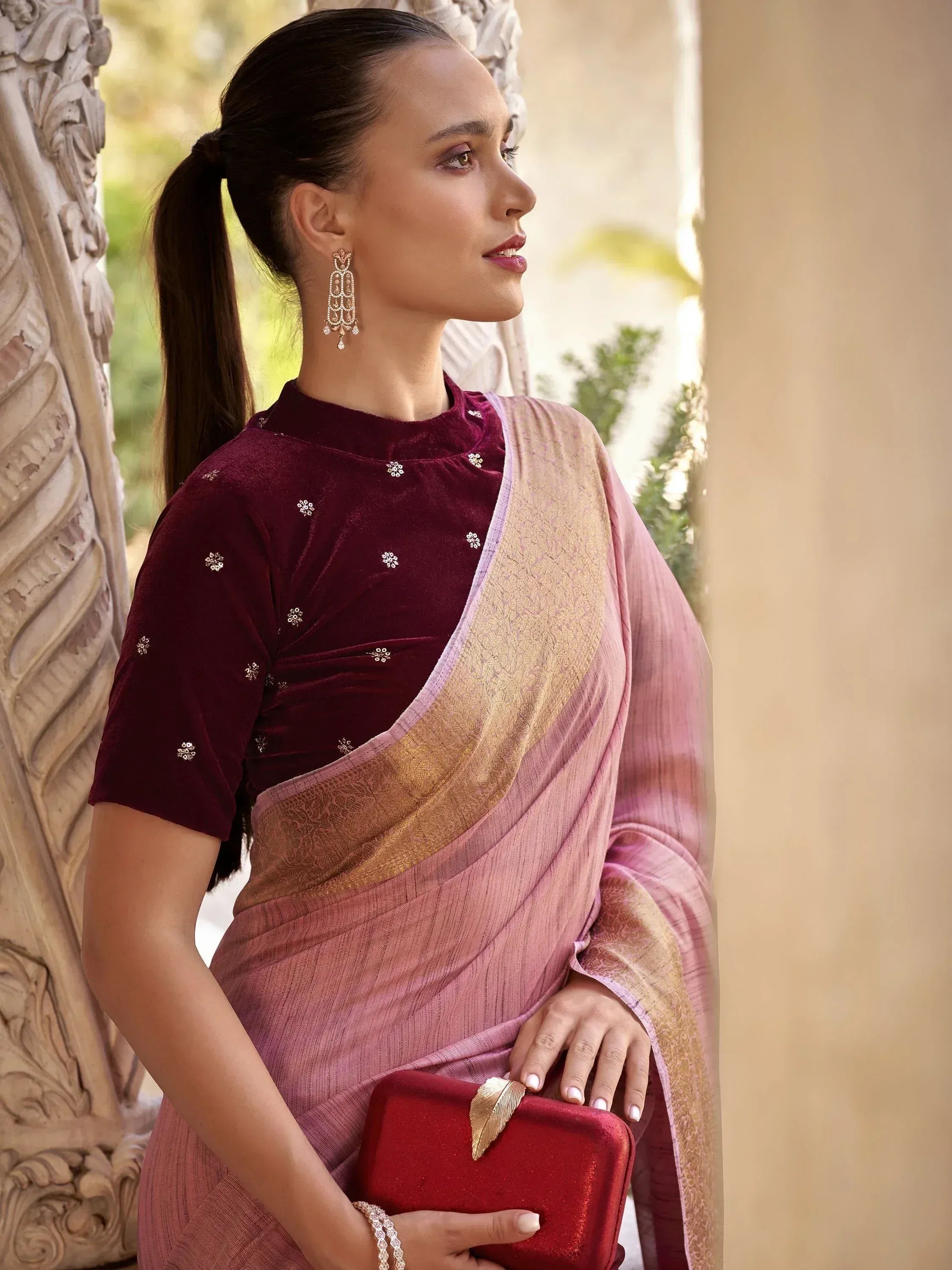 Woman in maroon velvet blouse with floral embroidery, pink saree with gold border, holding red clutch