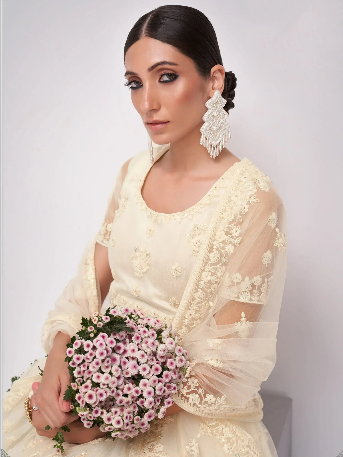Elegant ivory lehenga choli with floral embroidery, sheer sleeves, and statement earrings, holding pink flowers