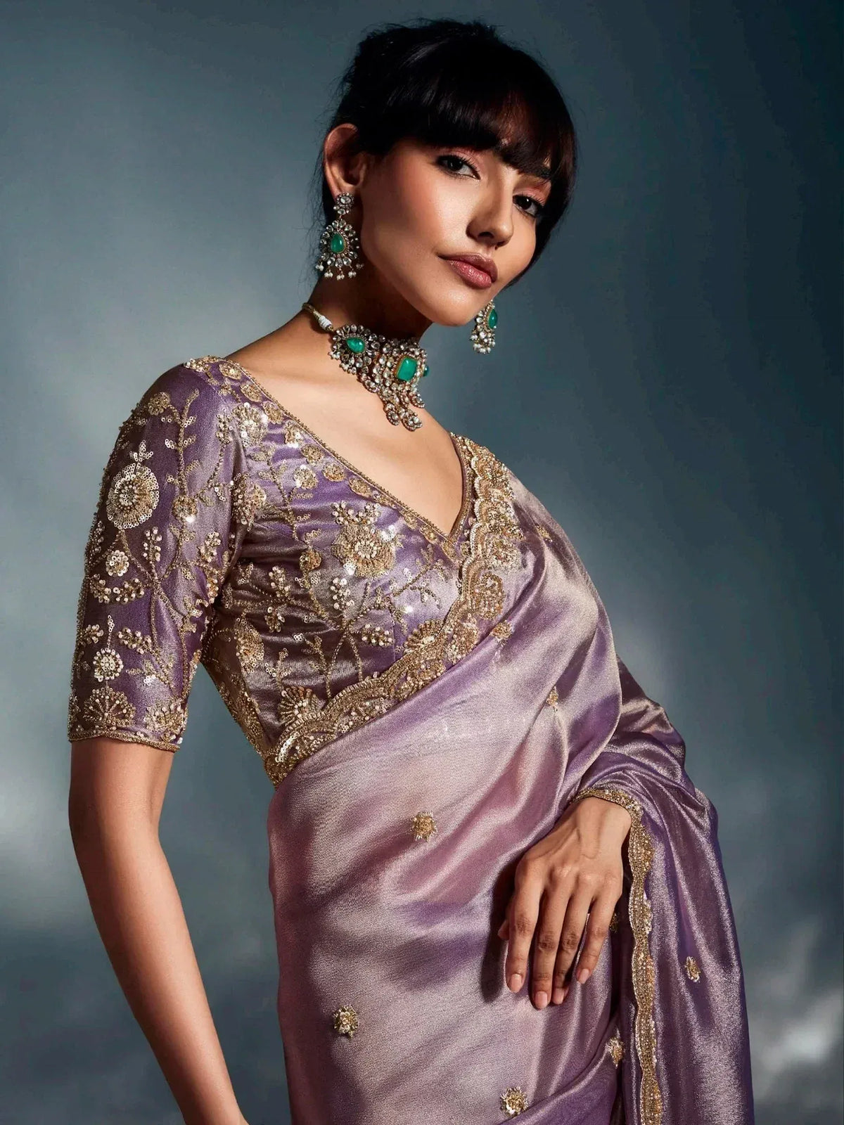 Lavender silk saree with gold floral embroidery and matching blouse, styled with emerald jewelry for Indian ethnic wear.