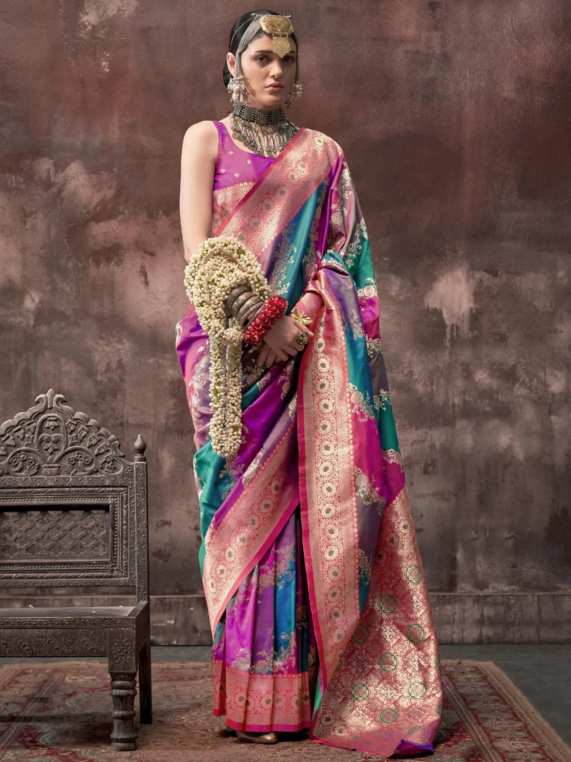 Woman in vibrant purple and teal silk saree with gold zari, traditional jewelry, and floral garland
