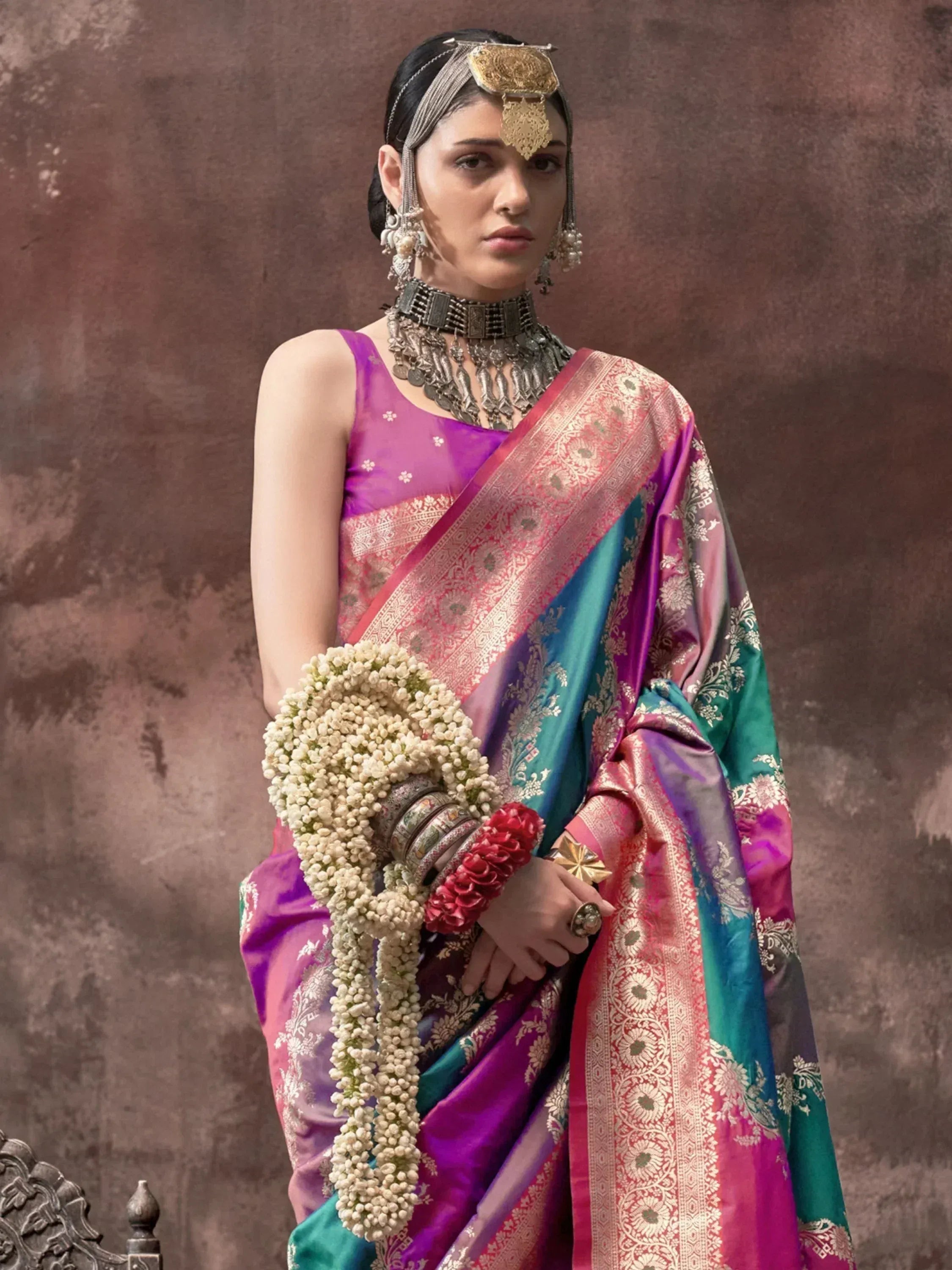 Woman in vibrant purple and teal designer saree with gold embroidery, traditional jewelry, and floral garland