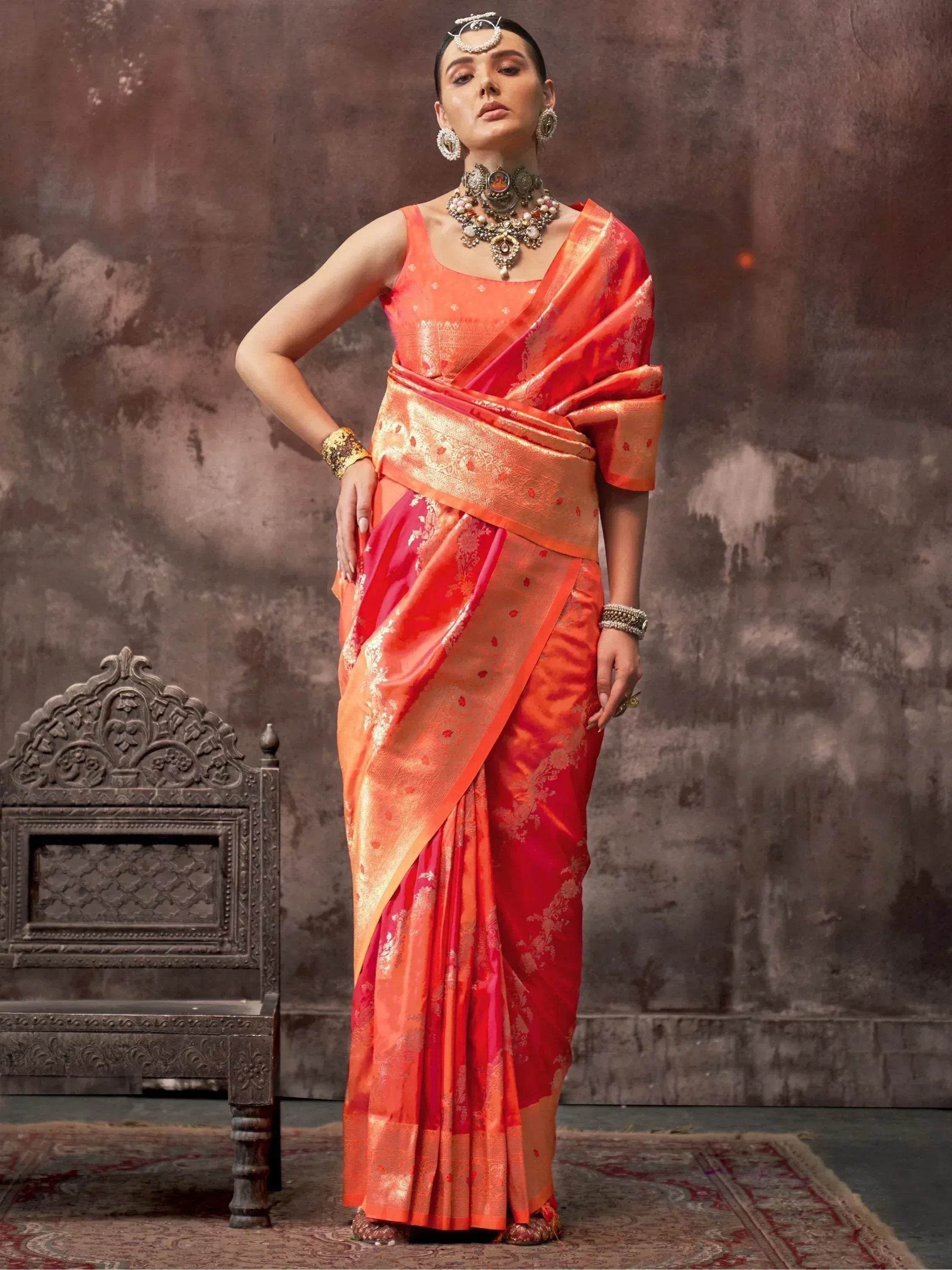 Woman in vibrant orange and red designer saree with gold embroidery, traditional jewelry, and ornate chair backdrop
