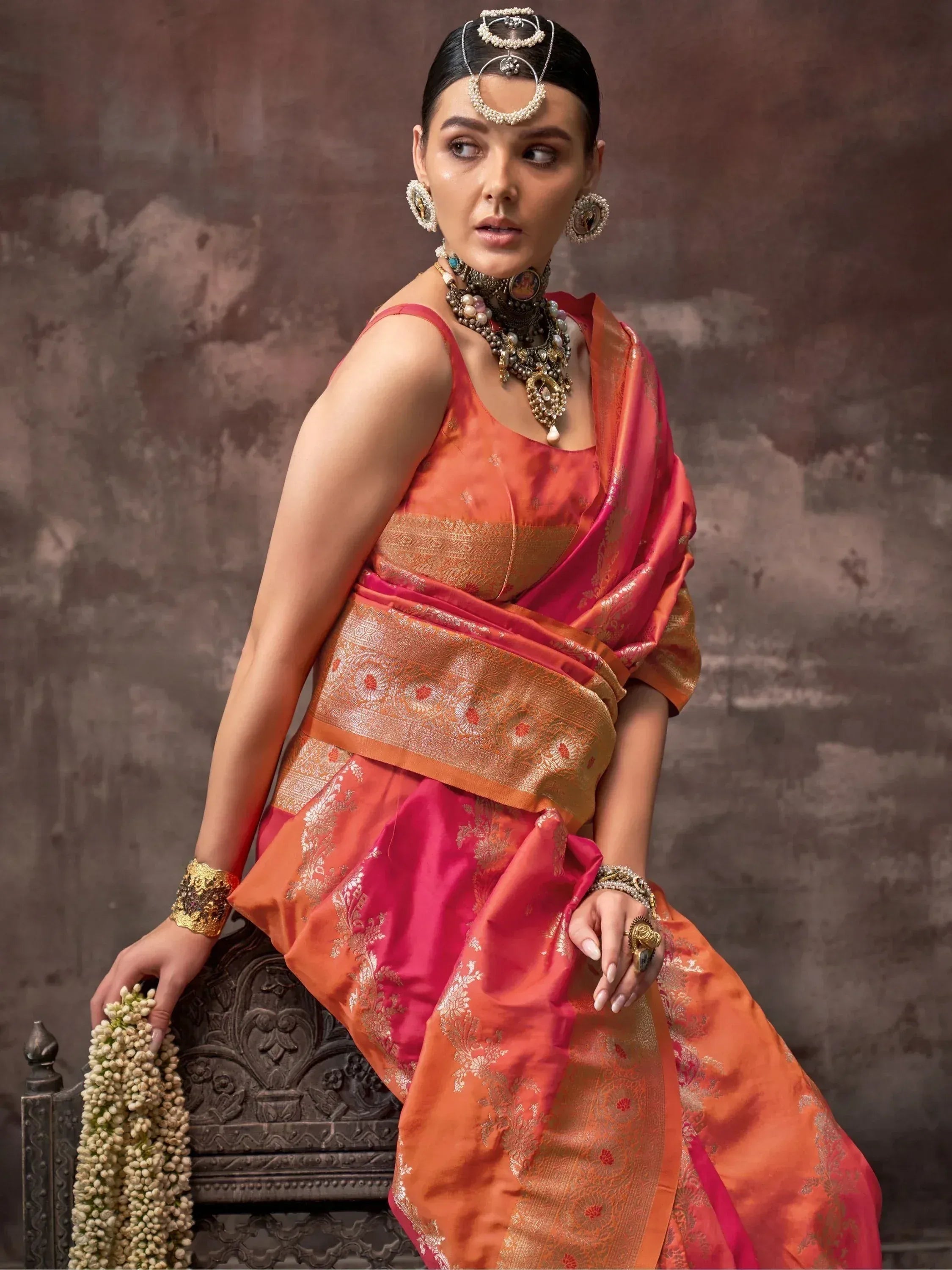 Woman in vibrant orange and pink designer saree with gold embroidery, traditional jewelry, and floral garland