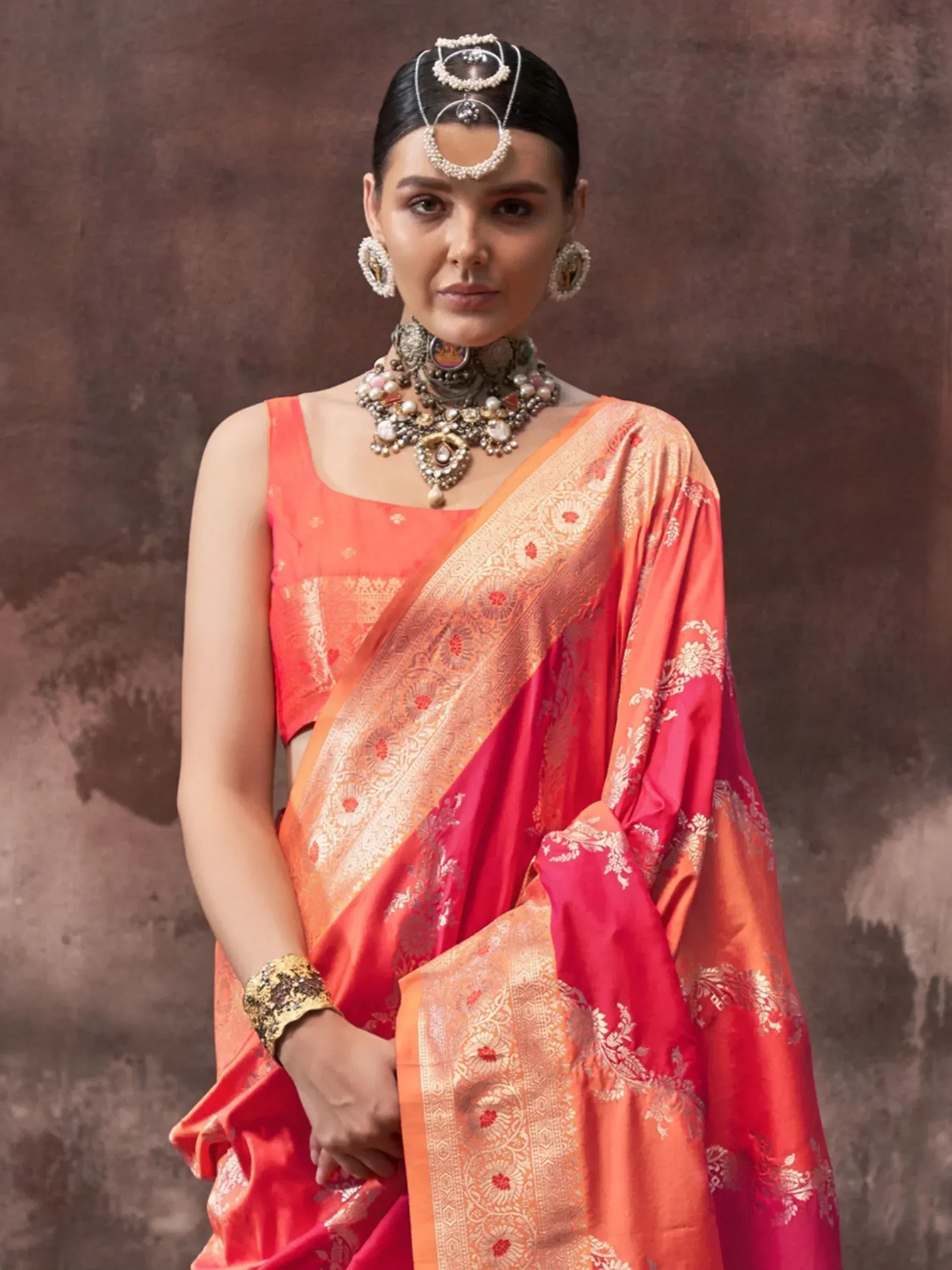 Woman in vibrant orange and pink designer saree with gold embroidery, traditional jewelry, and maang tikka